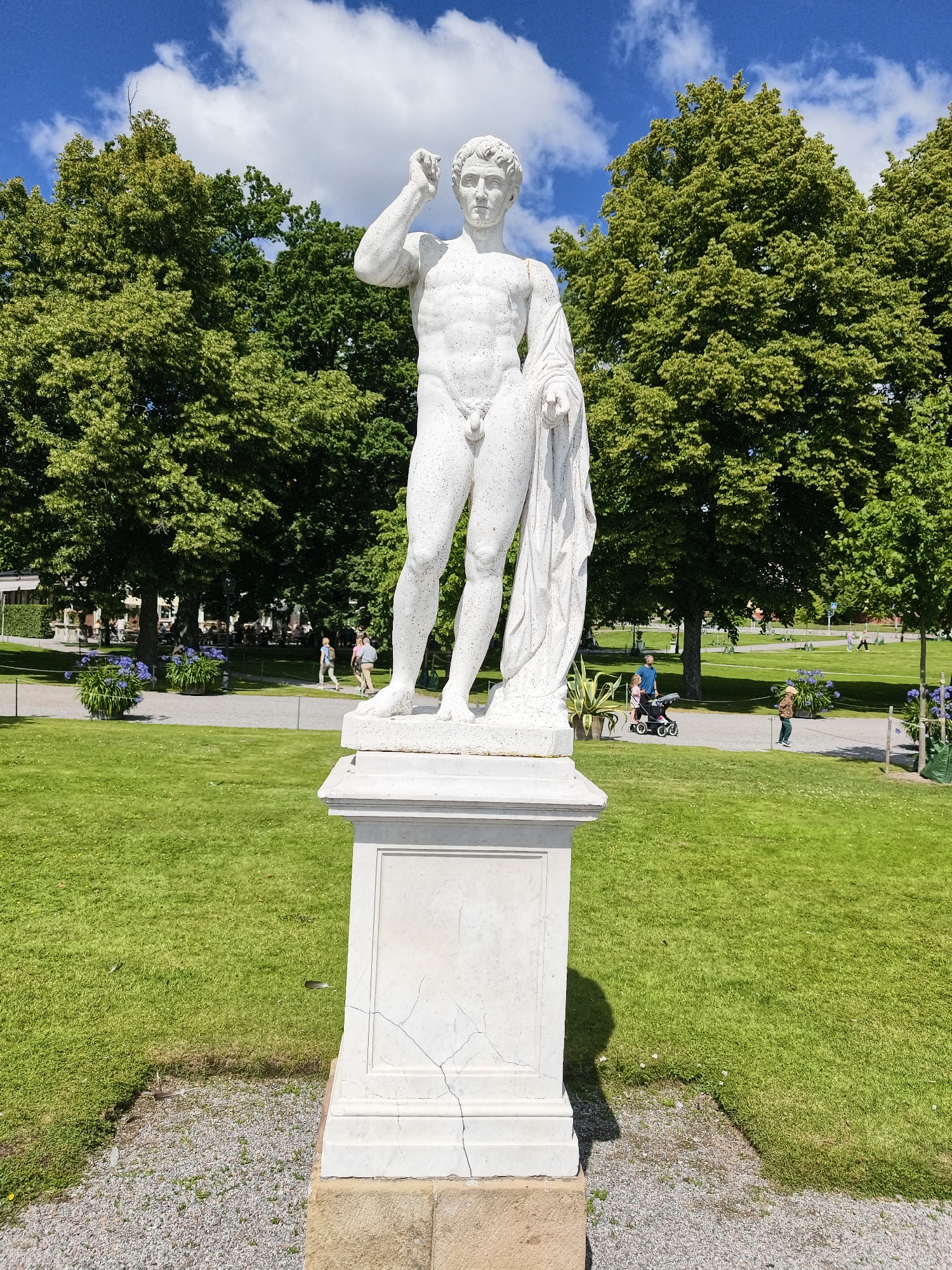 A white marble statue of a muscular, nude male figure stands on a pedestal in a sunlit park. The statue is draped with a white cloth over its left shoulder and arm, and its right arm is raised with the hand touching its forehead. The pedestal is white and shows visible cracks. Behind the statue, a large grassy lawn extends towards a line of mature green trees. In the middle ground, people are walking on a gravel path, some pushing strollers or with children. Further in the background, more trees and buildings are visible. The sky above is blue with scattered white clouds. The scene appears to be in a public garden or park in the city of Ekerö kommun, Sweden, likely during daytime.