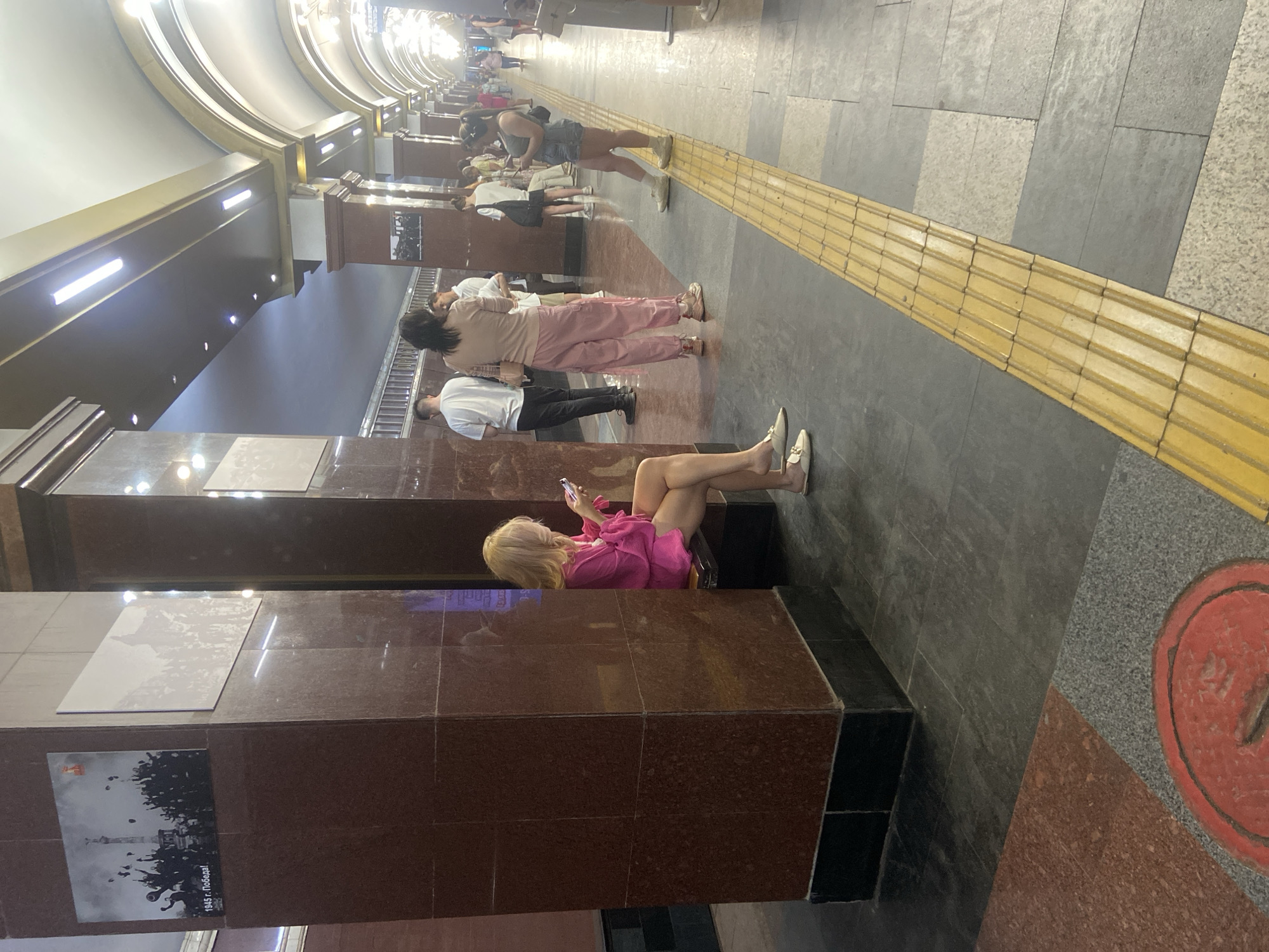 A view of a subway station platform with the walls covered with brown, granite slabs and decorative elements. Patrons wait or walk along the platform. A woman in a pink outfit sits on a ledge, using her phone. The station is located in Kazan, Russia.