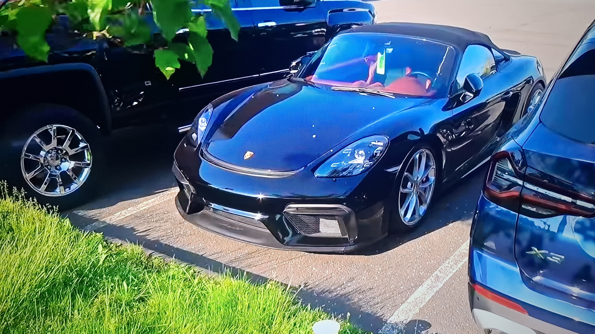 The image shows a dark blue Porsche 718 Spyder parked in a parking lot. The car has a red interior and aftermarket wheels. Next to the Porsche is a blue BMW X3. There is also a large black pickup truck in the background with chrome rims. The scene is an outdoor parking lot with some grass and trees.