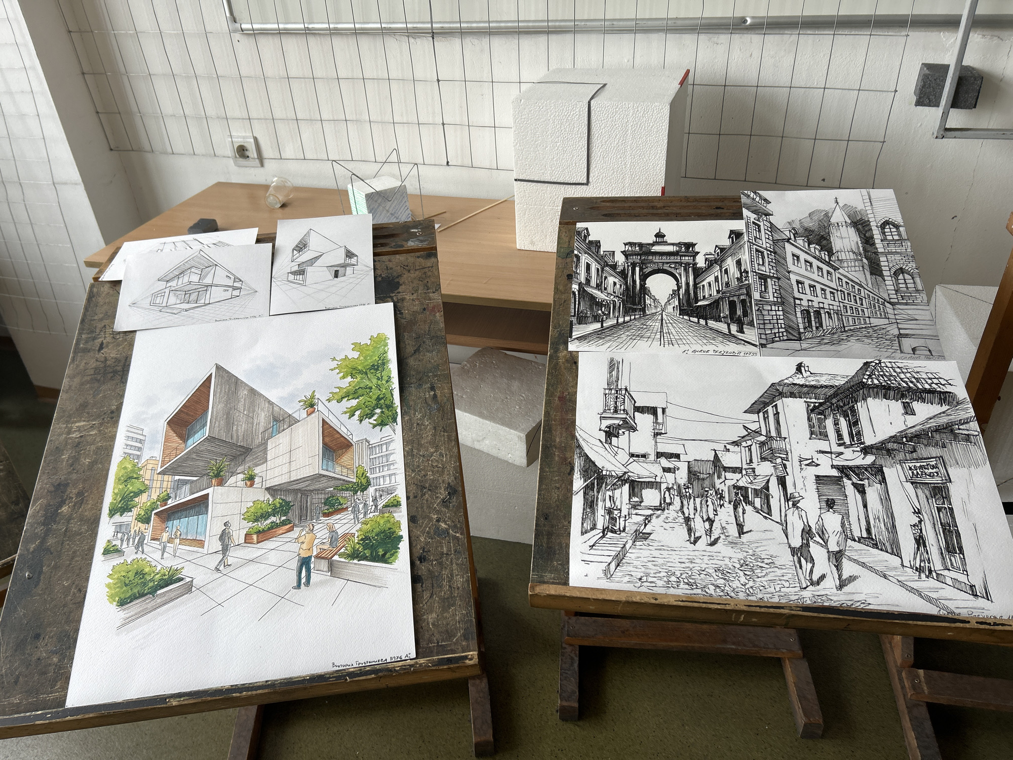This media file captures an indoor scene, likely an art or architecture studio in Sofia, Bulgaria, featuring two wooden easels displaying various drawings. The setting includes a wooden table against a white tiled wall with a metal grid. Several architectural models or abstract foam shapes are also visible on the table.
On the left easel, a large, detailed architectural rendering in color depicts a modern building with multiple levels, extensive landscaping, and several figures walking around. The style is contemporary, with clean lines and subtle shading. Above it, two smaller, black-and-white line drawings show perspective sketches of modern houses. The large drawing is signed "ВИКТОРИЯ ГРИГОРЬЕВА 1196 АГ" (Viktoria Grigorieva 1196 AG).
The right easel holds two black-and-white ink drawings. The top drawing presents a grand European street scene with an archway, historic buildings, and figures, signed "Р. АГНЕШКА 1195" (R. Agnieszka 1195). The bottom drawing depicts a narrower, older street with cobblestones, textured building facades, and several pedestrians. This drawing includes the Greek text "Ο ΚΟΣΜΟΣ" (The World) and another partially visible signature.
The scene suggests an academic or workshop environment where students' architectural and urban sketching works are being displayed or reviewed. The time of day appears to be daytime, with general ambient light. A small, empty glass bottle and a dark object are on the table.