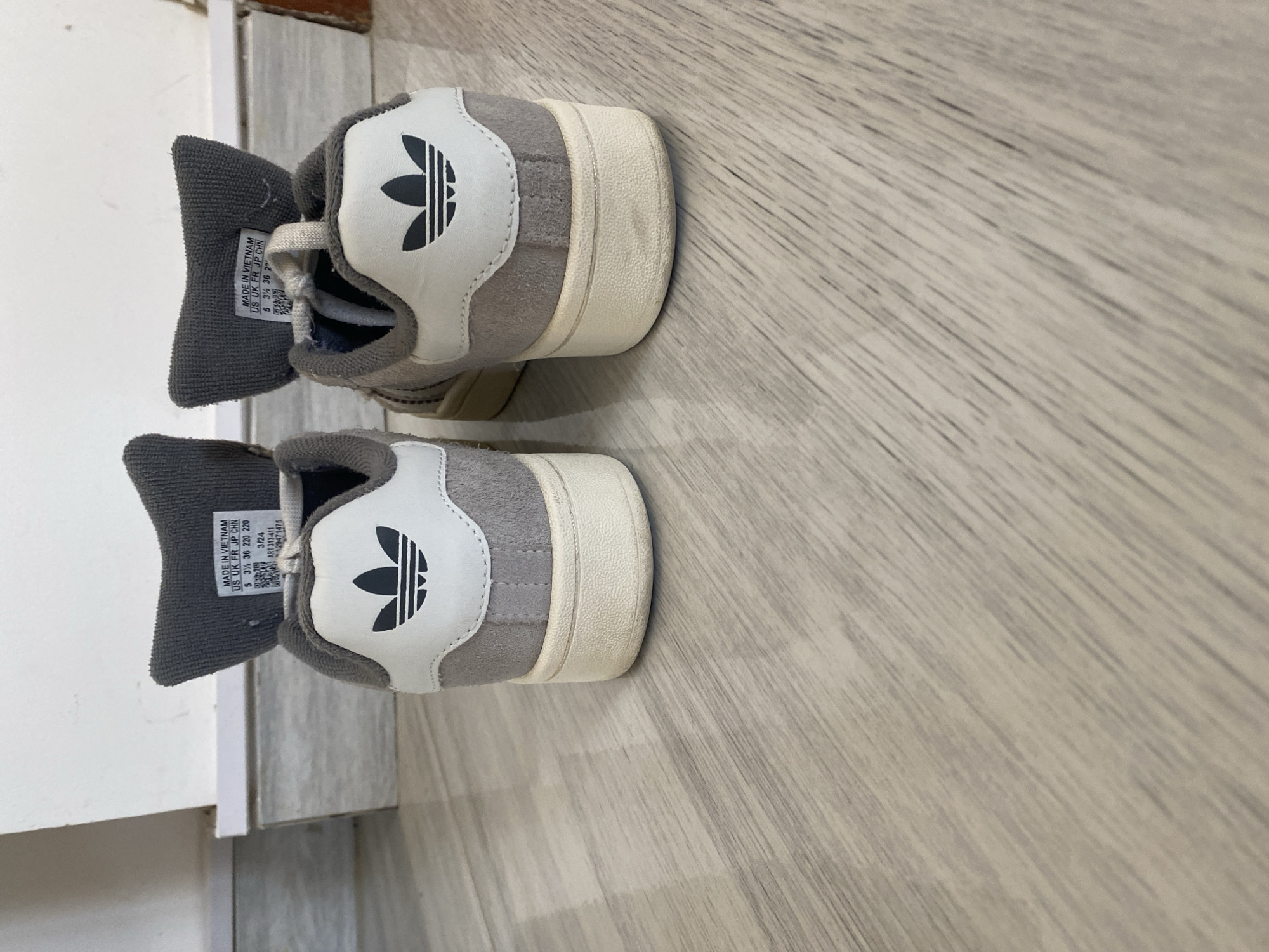 The image shows the back view of two Adidas sneakers on a wooden floor. The sneakers are light gray with white accents and feature the Adidas logo on the heel tab. The floor has a light wood grain pattern. The background is out of focus but appears to be a wall and a baseboard. There are no people or specific landmarks visible to suggest a location in Sharm El Sheikh, Egypt.
