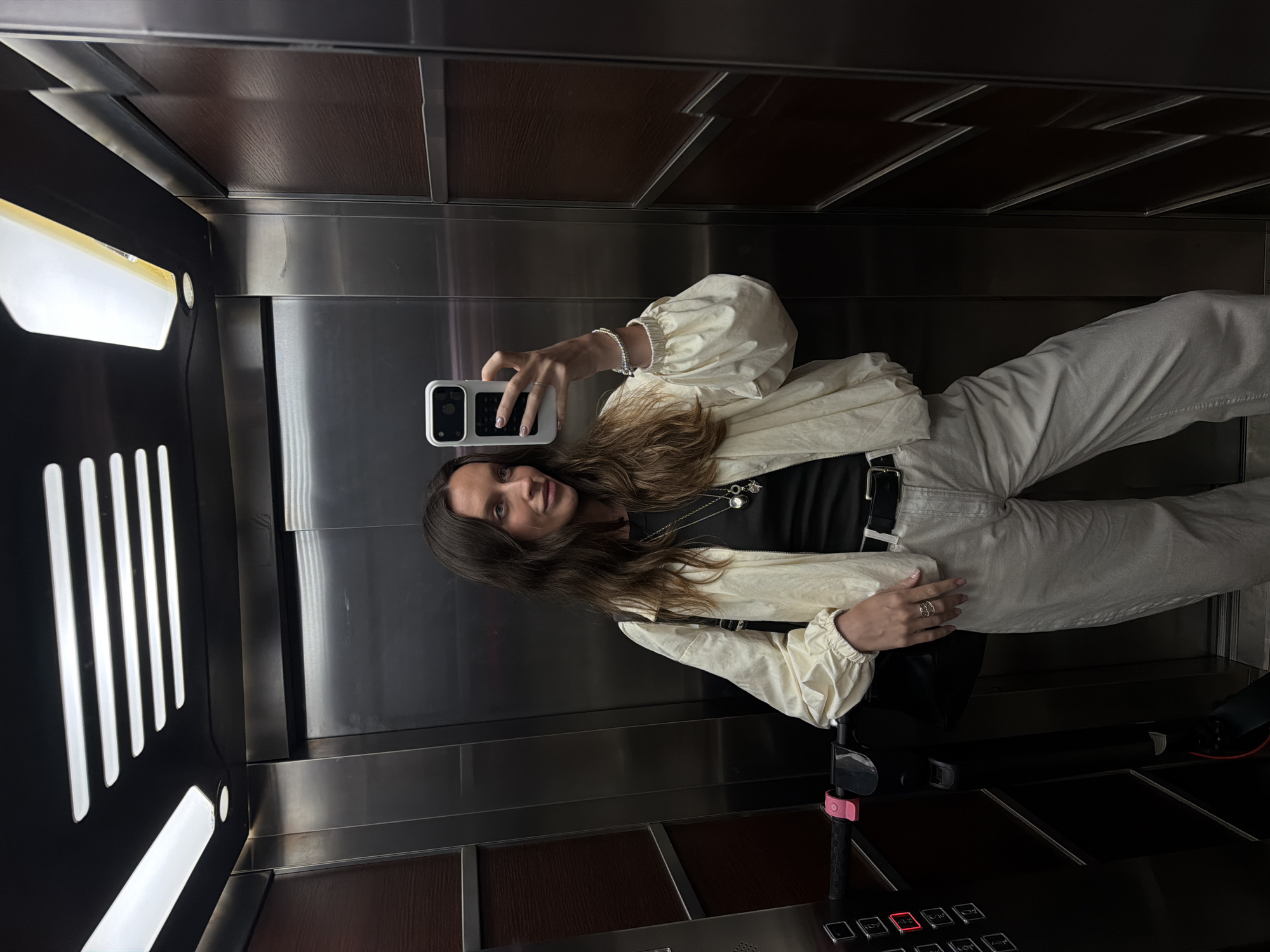 A young woman with long, brown hair is taking a selfie in an elevator. She is wearing a black t-shirt, a cream-colored bomber jacket with puffed sleeves, and light-colored pants. She has a black belt and is wearing jewelry, including necklaces and bracelets. She is holding a white phone in her right hand. To her left, there is a black electric scooter with a pink brake lever. The elevator has stainless steel walls and wooden panels. The ceiling has fluorescent lighting. The image appears to have been taken in Budva, Montenegro.
