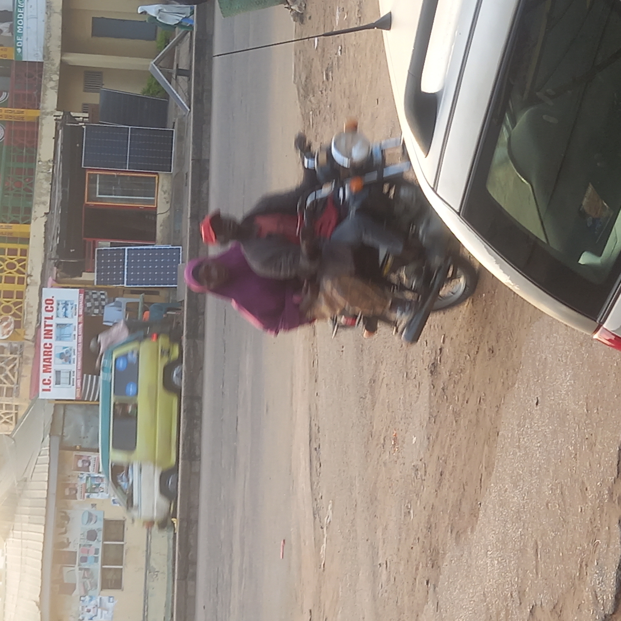 The image shows a street scene in Sardauna, Nigeria. A motorcycle with two riders is traveling down the road, next to a white car. A yellow van is visible near a building that has solar panels displayed. The building also has a sign that reads "I.C. MARC INT'L CO." The road is dirt.