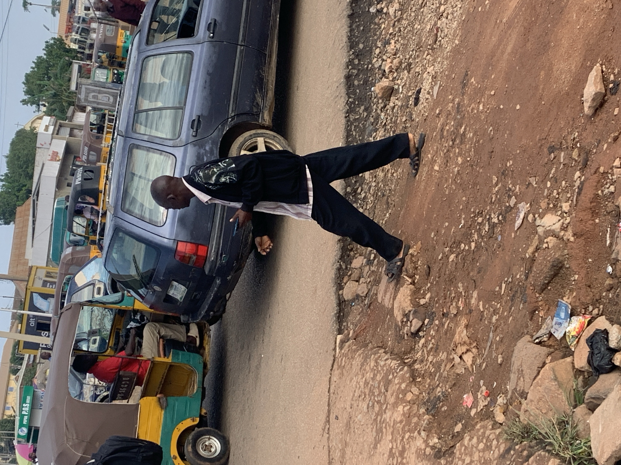 The image depicts a street scene in the city of Gura Top, Nigeria, characterized by a dusty, unpaved road with scattered debris and rocks. In the foreground, a man is walking, wearing dark pants and a patterned jacket over a striped shirt, with dark sandals on his feet. He is holding a mobile phone in his right hand and appears to be looking down at it.
Behind him, a dark-colored station wagon is parked, partially obscuring the view of other vehicles. To the left of the station wagon, a yellow and green auto-rickshaw, also known as a "keke," is visible. A man wearing a red shirt is seated in the driver's seat of the keke. Further in the background, several other auto-rickshaws and various buildings and structures are discernible, suggesting a commercial or busy urban area. The overall lighting indicates it is daytime, with a slightly overcast or hazy sky. The environment appears to be a typical roadside setting in a Nigerian town, with elements of local transportation and urban infrastructure.