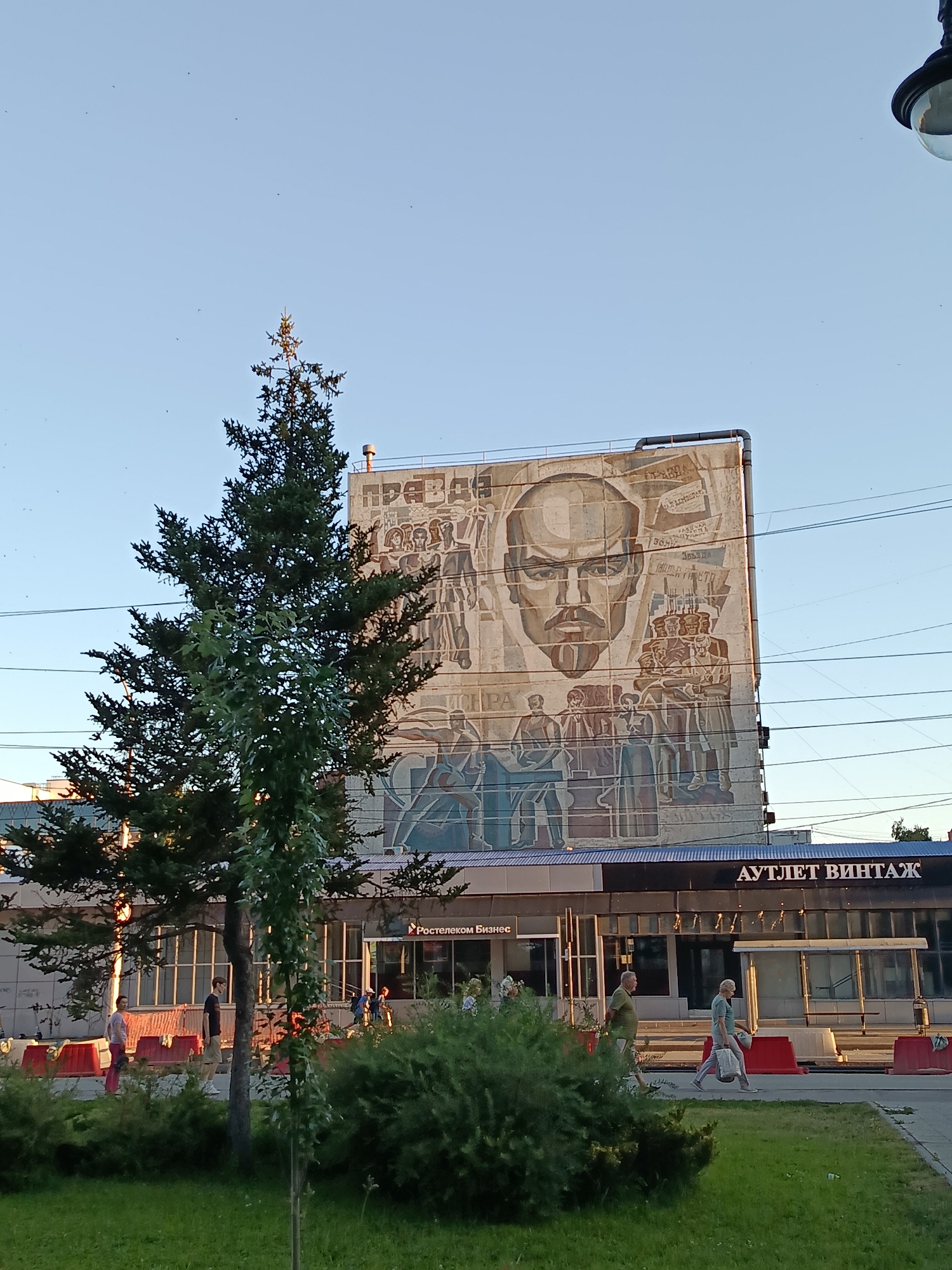 The image shows a building in Saratov, Russia, adorned with a large mural. The mural depicts a portrait of Vladimir Lenin and scenes of collective labor and ideological messages in Russian. Below the mural, the building has a storefront with signs reading "Ростелеком Бизнес" (Rostelecom Business) and "АУТЛЕТ ВИНТАЖ" (Outlet Vintage). A tall evergreen tree stands in front of the building, partially obscuring the mural. The area in front of the store features a sidewalk, grass, and red temporary barriers. Several people are visible walking along the sidewalk. The sky is clear and blue, indicating it is daytime. Overhead power lines are also visible.