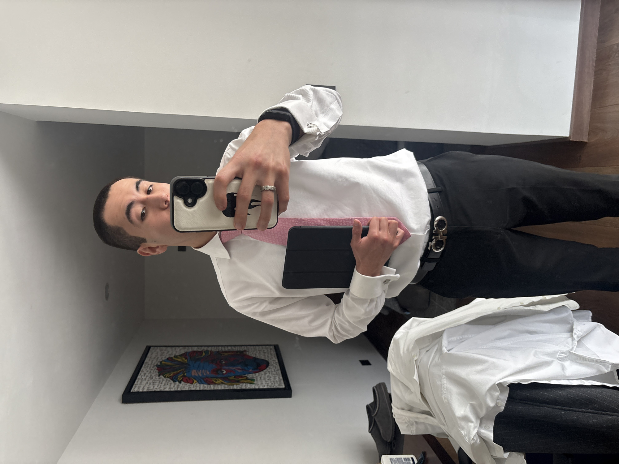 A person is taking a selfie in what appears to be a bathroom or dressing area. The person has short, dark hair and is wearing a white, long-sleeved dress shirt, a light pink tie, and black pants with a black belt and a silver buckle. The person is holding a smartphone in their left hand with a phone case. They are also holding a black tablet in front of them. The background includes a white wall, a framed piece of artwork on the wall depicting a face with colorful details, a glimpse of dark wood flooring, and some clothing items including a white dress shirt and dark colored pants draped over what appears to be a chair or other furniture. The specific location in San Bernardino Tlaxcalancingo, Mexico, cannot be determined from the visual information provided.