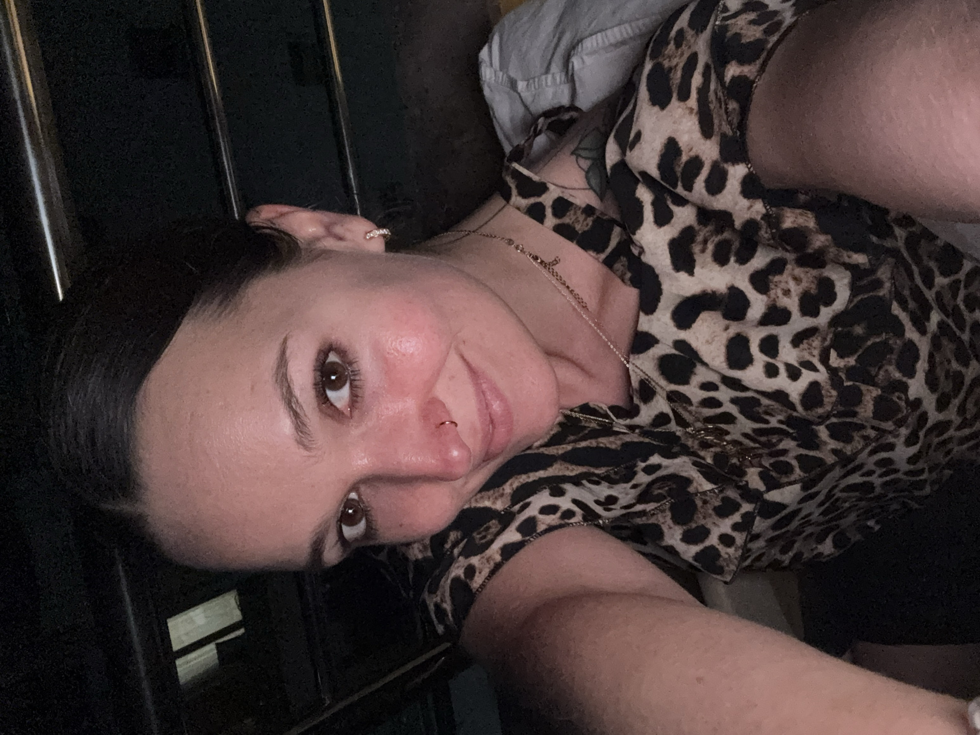 In an indoor setting, possibly a vehicle at night, a woman with dark hair pulled back is taking a selfie. She is wearing a leopard-print top with a gold necklace featuring a cross charm. She has a nose ring and small hoop earrings. Her skin appears flushed, and she is smiling faintly. The background is dark, with hints of metallic railings or bars above her head, and a rectangular illuminated object to her left. The scene does not provide specific details to pinpoint a location within Budva, Montenegro.