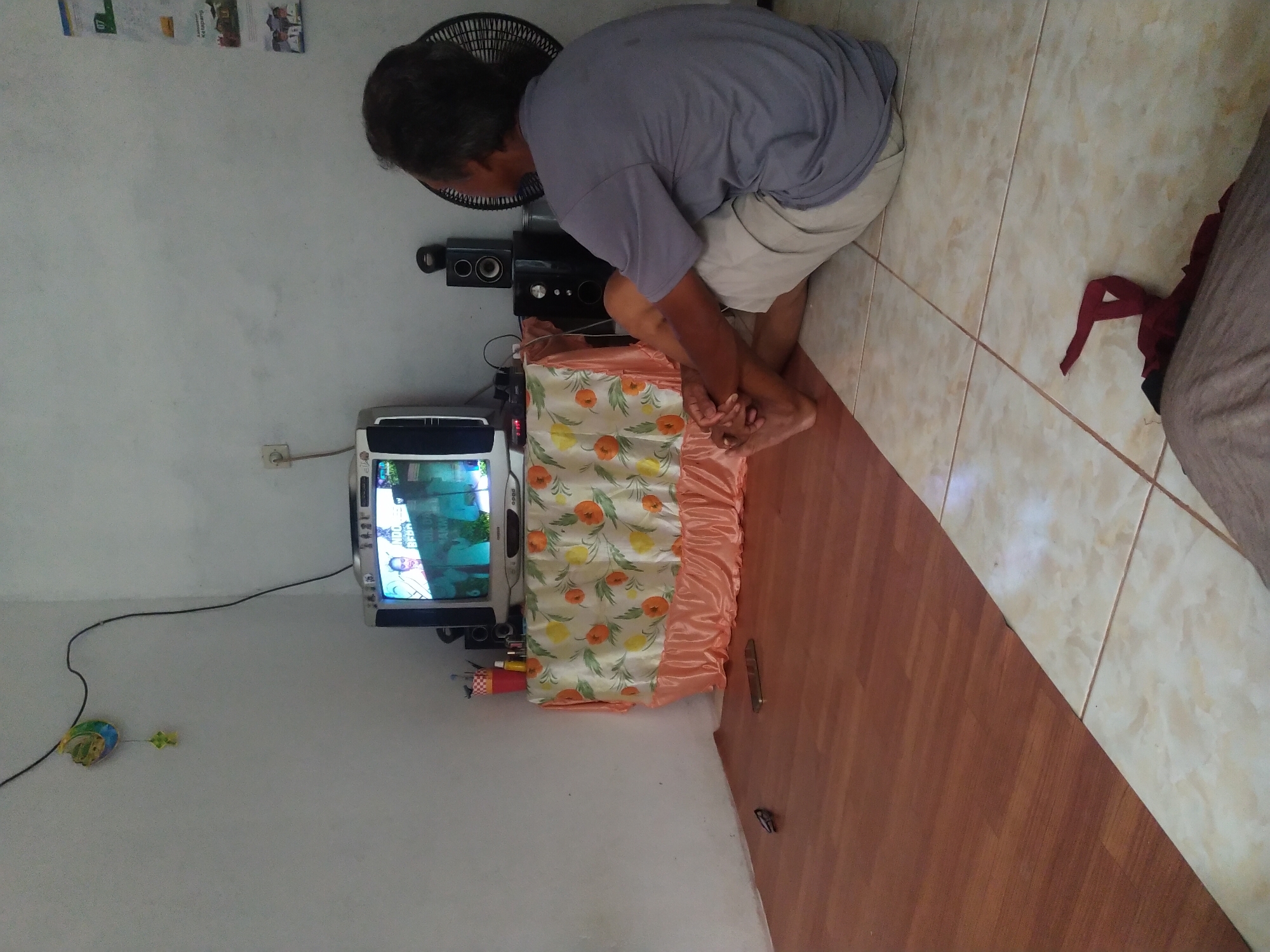 A medium shot captures an elderly Indonesian man sitting on the floor in front of a television in a modest room. The man, with graying hair, is wearing a gray t-shirt and light-colored shorts. His legs are crossed, and his hands are clasped between his knees, indicating a relaxed posture as he watches the television.
The television set, an older model with a curved screen, displays a program featuring a woman. The TV is placed on a small table covered with a fabric adorned with orange and yellow fruit patterns and a ruffled, peach-colored trim. To the right of the TV, there are two black speakers and a black oscillating fan. On the wall above the television, a string of decorative items, including a colorful kite-like object, hangs from a black wire. A power outlet is visible on the wall to the left of the television.
The room has two distinct floor coverings: a wood-like laminate floor in the foreground and a tiled floor in the background where the man is seated. A dark red piece of cloth is on the tiled floor in the lower right corner. On the wood floor near the television, a pair of eyeglasses and a mobile phone are present. The walls are painted white, and some posters or papers are affixed to the wall on the right side of the image, suggesting it might be a home or a small office. The scene appears to be within a dwelling in Sintang, Indonesia, given the context provided.