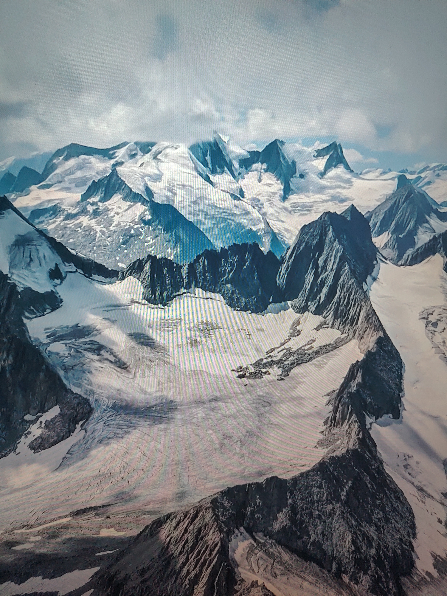 The image depicts a mountainous landscape covered in snow and glaciers under a cloudy sky. The foreground features dark, rocky mountain ridges. Glaciers and snowfields fill the valleys between the mountains. The location is unknown.