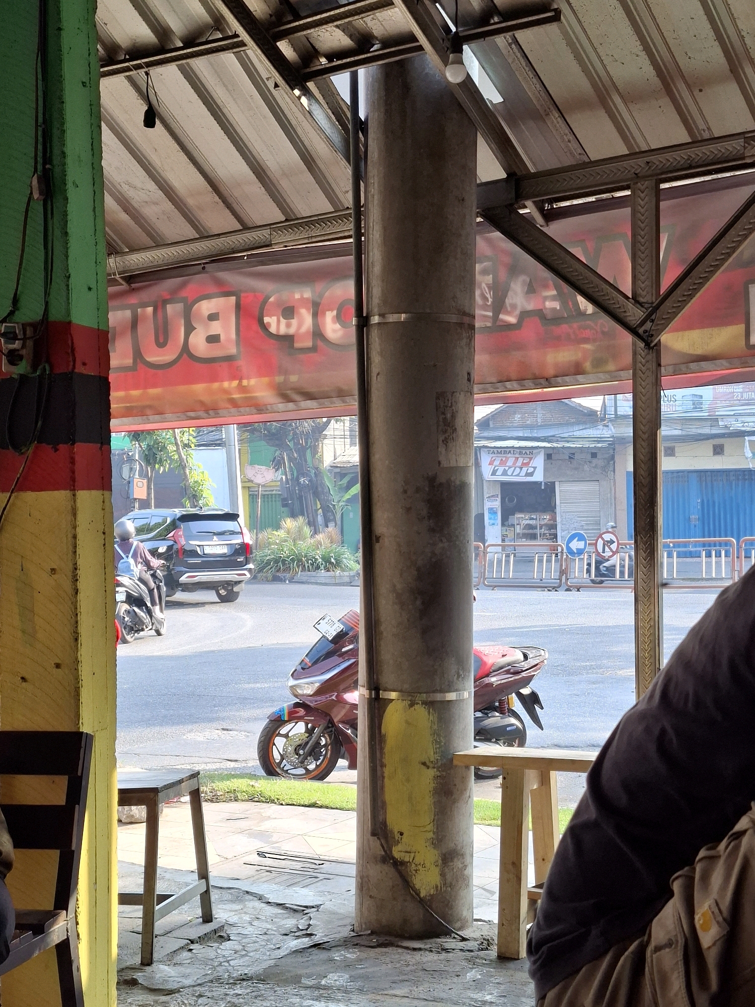 The image captures a street-level view from beneath a covered structure, likely an awning or part of a building, looking out onto a street in Surabaya, Indonesia. In the foreground, a thick, gray metal pillar stands prominently, with faded yellow paint marks near its base. To the left, a green-painted support column with red and black stripes is visible, with electrical wires attached. A wooden chair and table are partially seen on the left, and another small wooden table is on the right.
Beyond the pillar, the street is bustling with activity. A dark-colored SUV is seen from the rear, turning on the road, with a motorcycle rider behind it. Further down the street, various buildings line the road, including a shop with a roller shutter door and a sign that reads "TAMBAL BAN" (tire repair) with "TIP TOP" written on it. Traffic signs, including a blue left-arrow sign and a red circle with a diagonal line and arrow, are present on the sidewalk. Several metal barriers are also visible along the roadside.
The background features more buildings and greenery, suggesting a typical urban streetscape. The overall lighting indicates it is daytime, with bright sunlight casting shadows. The image appears to be taken from a sheltered area, possibly a roadside eatery or shop, looking out into the street.