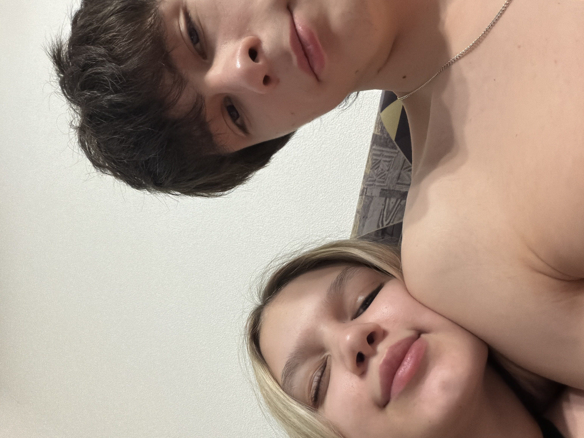 A close-up, selfie-style photograph features two young individuals, a female and a male, in an intimate pose. The female, positioned on the left, has blonde hair and fair skin, with her eyes closed or nearly closed, and a subtle smile on her lips. Her head rests gently against the male's right shoulder/upper chest. The male, positioned on the right, has dark, somewhat disheveled hair and fair skin, looking directly at the camera with a neutral expression. Both individuals appear to be shirtless, exposing their bare shoulders and chests. The male wears a thin silver chain necklace around his neck. Behind them, a light-colored, possibly white or off-white, textured wall serves as the primary background. To the lower right, partially visible behind the male, is a section of patterned fabric or upholstery, displaying a geometric design in muted tones, possibly brown, grey, and yellow. The lighting is soft and even, suggesting an indoor setting. The specific location within the city of Yekaterinburg, Russia, cannot be determined from the visual information present in the image.