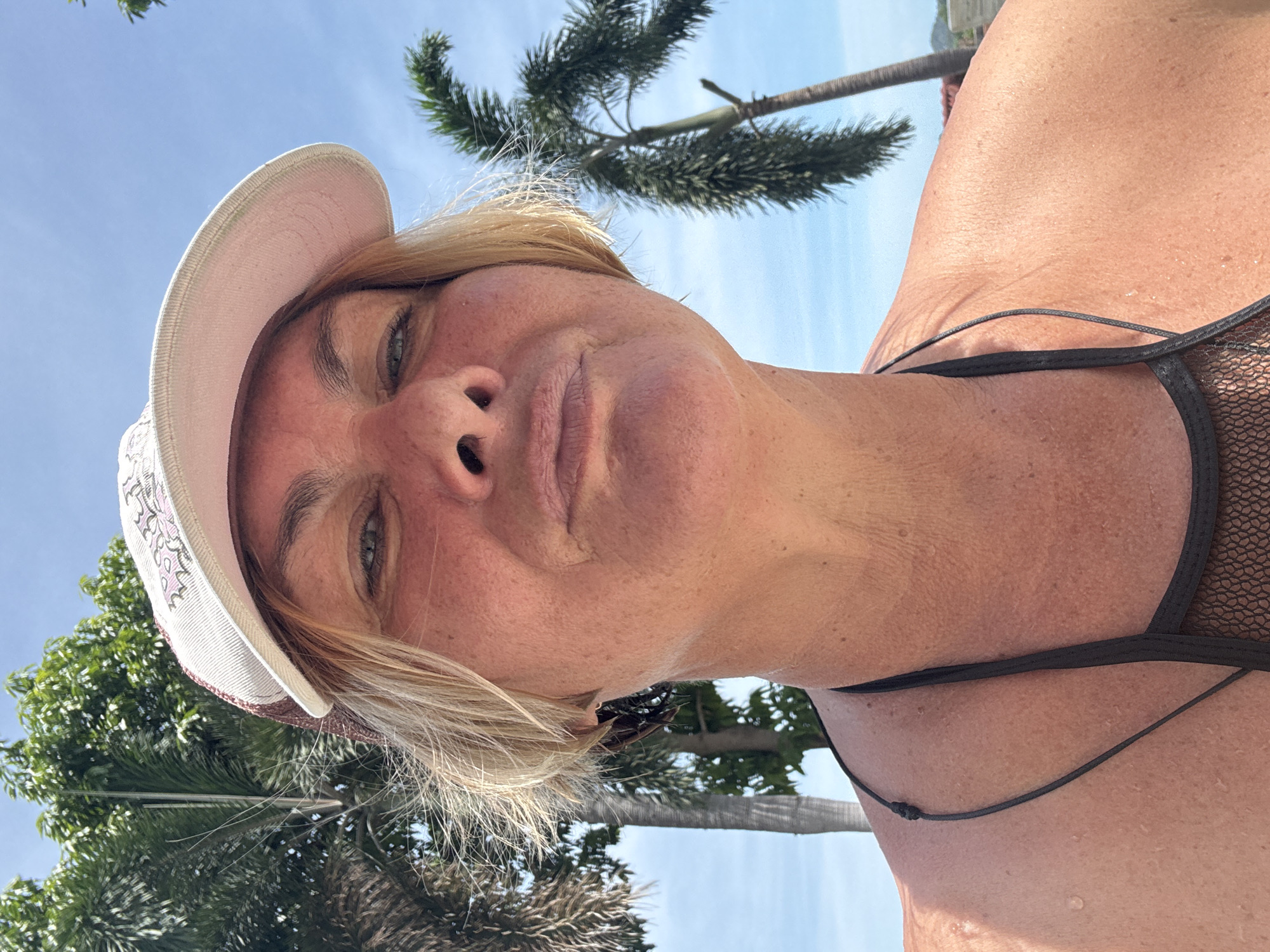 A sunlit close-up portrait shows a fair-skinned woman wearing a white cap with floral embroidery and a black bikini top. Her blonde hair is visible beneath the cap. The woman's face is oriented at an upward angle towards the camera. The background features tropical foliage, likely palm trees, against a blue sky. The location is บ้านบางรักษ์, Thailand.
