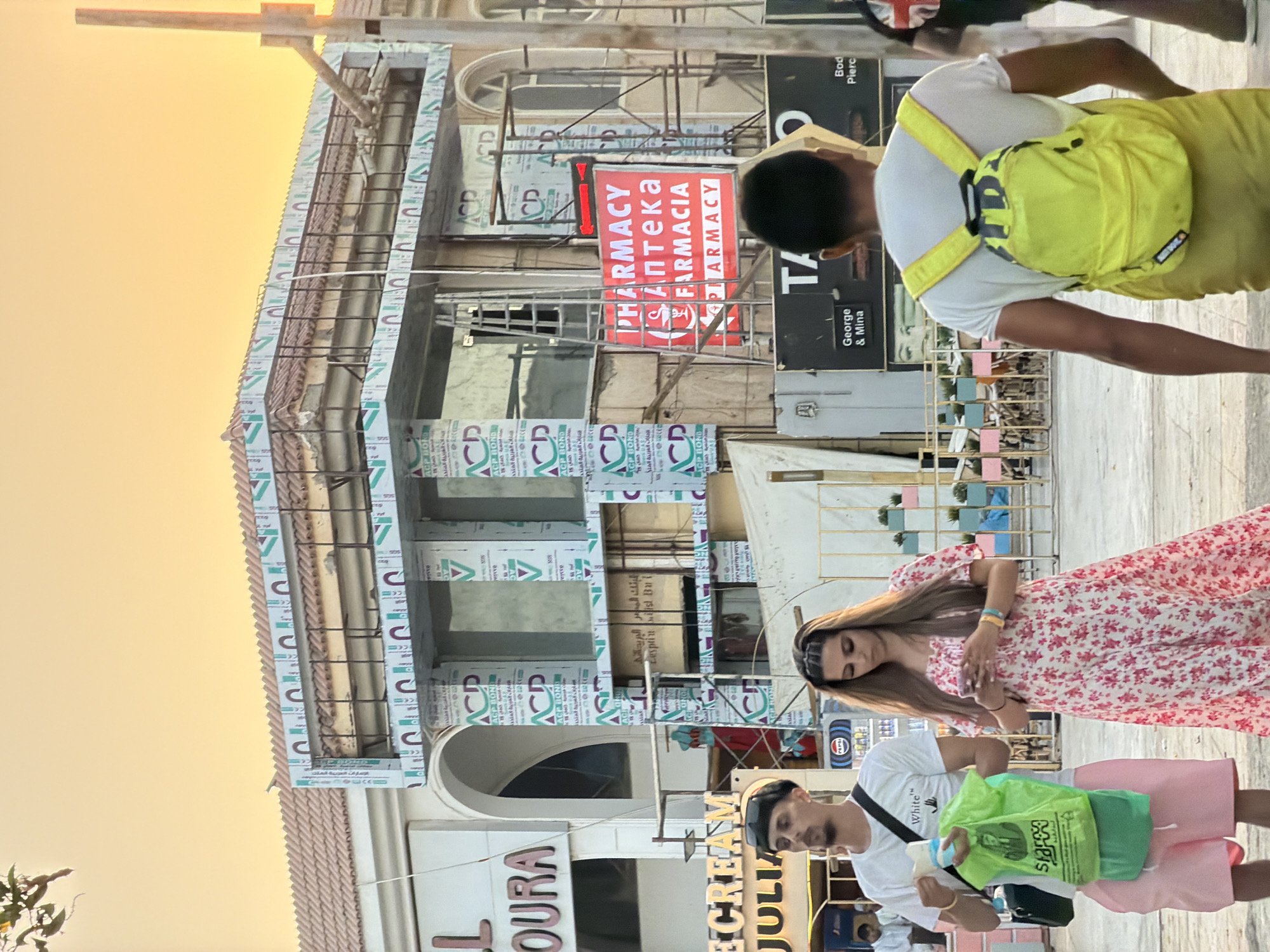 In Sharm El Sheikh, Egypt, an outdoor scene unfolds with a building under construction in the background, adorned with panels featuring Arabic script and the logo "ACP BOND". A bright red illuminated sign displays "PHARMACY", "ΑΠΤΕΚΑ", and "FARMACIA" in multiple languages, indicating a pharmacy. In the foreground, a man wearing a black beanie, white t-shirt, and pink shorts walks with a green shopping bag and a black crossbody bag. To his right, a woman in a pink floral dress looks at her phone. Further in the background, a person with a bright yellow backpack walks away from the camera. The environment suggests a public street or promenade with cafes or shops visible behind the construction. The sky is a warm, hazy orange, indicative of sunset or sunrise.