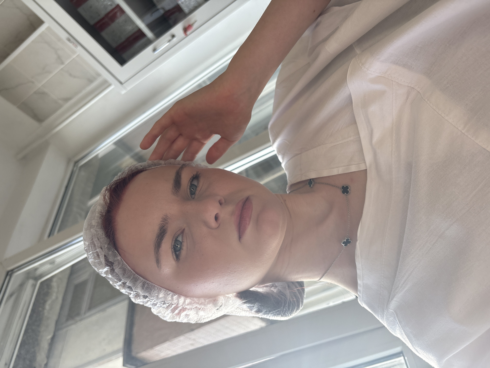 A young Caucasian female with red hair, wearing a white disposable hair net and a white robe, is posing for a selfie. She is indoors, with sunlight streaming in from a window on the left side of the frame. The background shows a window with a view of an urban environment, and a white cabinet with shelves containing various items. The woman's hand is raised to her head, and she is looking directly at the camera with a neutral expression. She is wearing a silver necklace with black clover-shaped pendants. The setting appears to be a salon, spa, or medical facility.