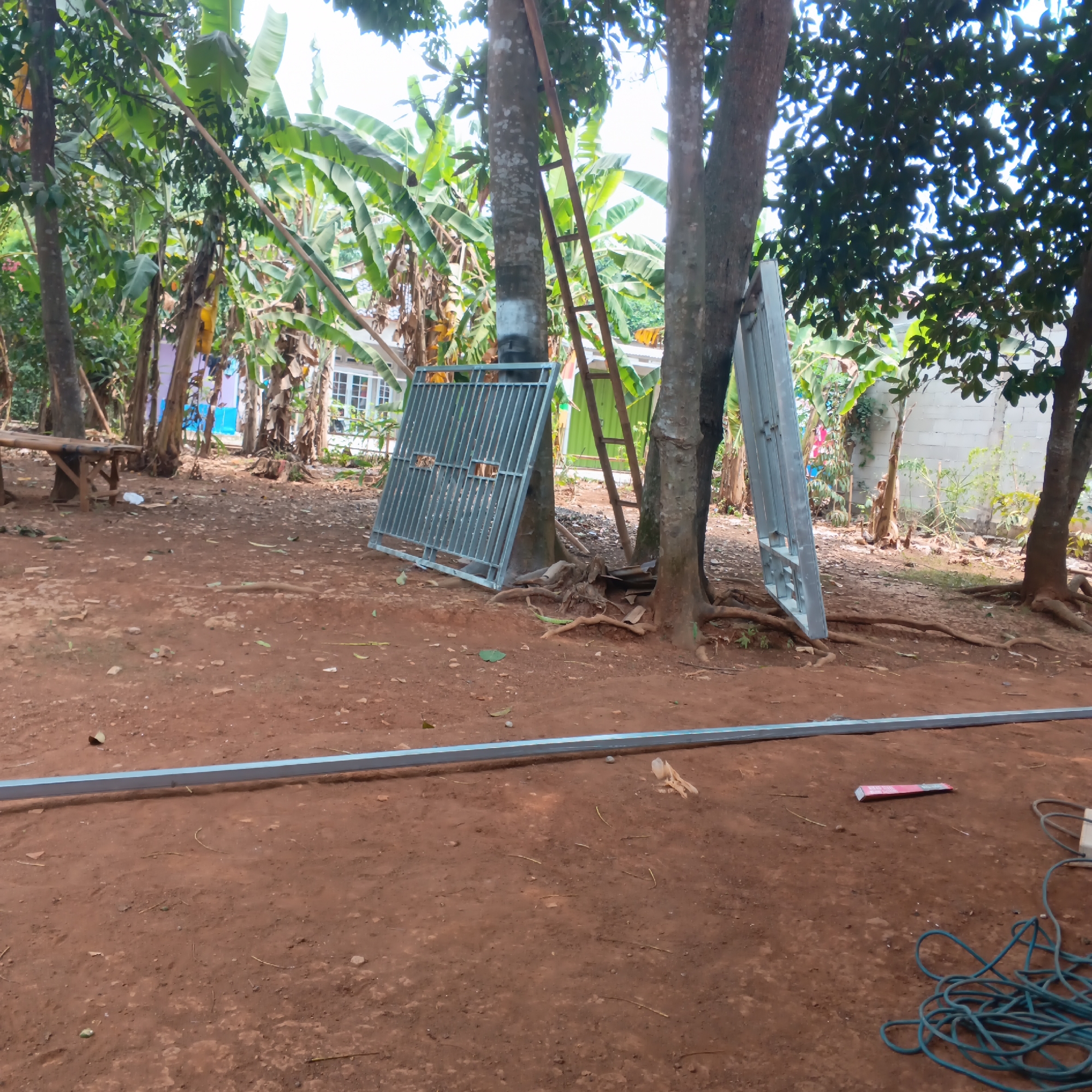 The image depicts an outdoor scene in Jayasampurna, Indonesia, likely a residential yard or an area undergoing construction. The ground is predominantly red-brown soil, scattered with small stones and dry leaves. In the foreground, a long, silver metal pipe lies horizontally on the ground. To the right, a coiled blue electrical cord and a small white box are visible.
In the midground, several trees with visible root systems are present. Leaning against one of these trees is a wooden ladder. Two large, gray metal gate frames are also prominent; one is leaning upright against a tree, and the other lies flat on the ground in front of it. Behind these objects, banana trees with large leaves and other foliage are visible, suggesting a tropical or subtropical environment.
In the background, a building with white walls and multiple windows is partially obscured by trees and vegetation. The overall impression is a somewhat cluttered outdoor space, possibly in a semi-rural or suburban setting, with elements of both natural vegetation and construction materials. No people are visible in the image.