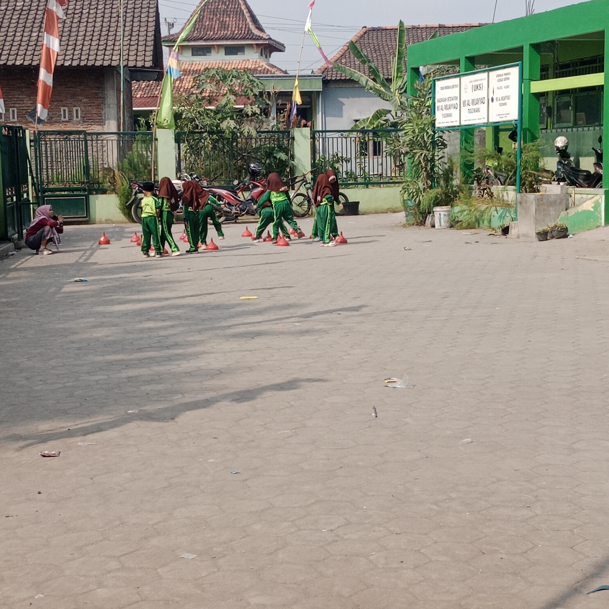 In Tegowanu Kulon, Indonesia, a group of children in green uniforms are engaged in a physical activity on a paved outdoor area. They are positioned around red cone markers, suggesting an obstacle course or drill. One child is further back in the scene, and another individual, a woman wearing a headscarf and maroon clothing, is crouched to the left, seemingly observing or photographing the activity. The environment features a building with a bright green facade and a roof with clay tiles. A fence runs along the front of the property, and several motorcycles are parked nearby. The ground is paved with interlocking bricks, and there are plants and trees scattered throughout the area. The overall impression is of an outdoor educational or recreational space.