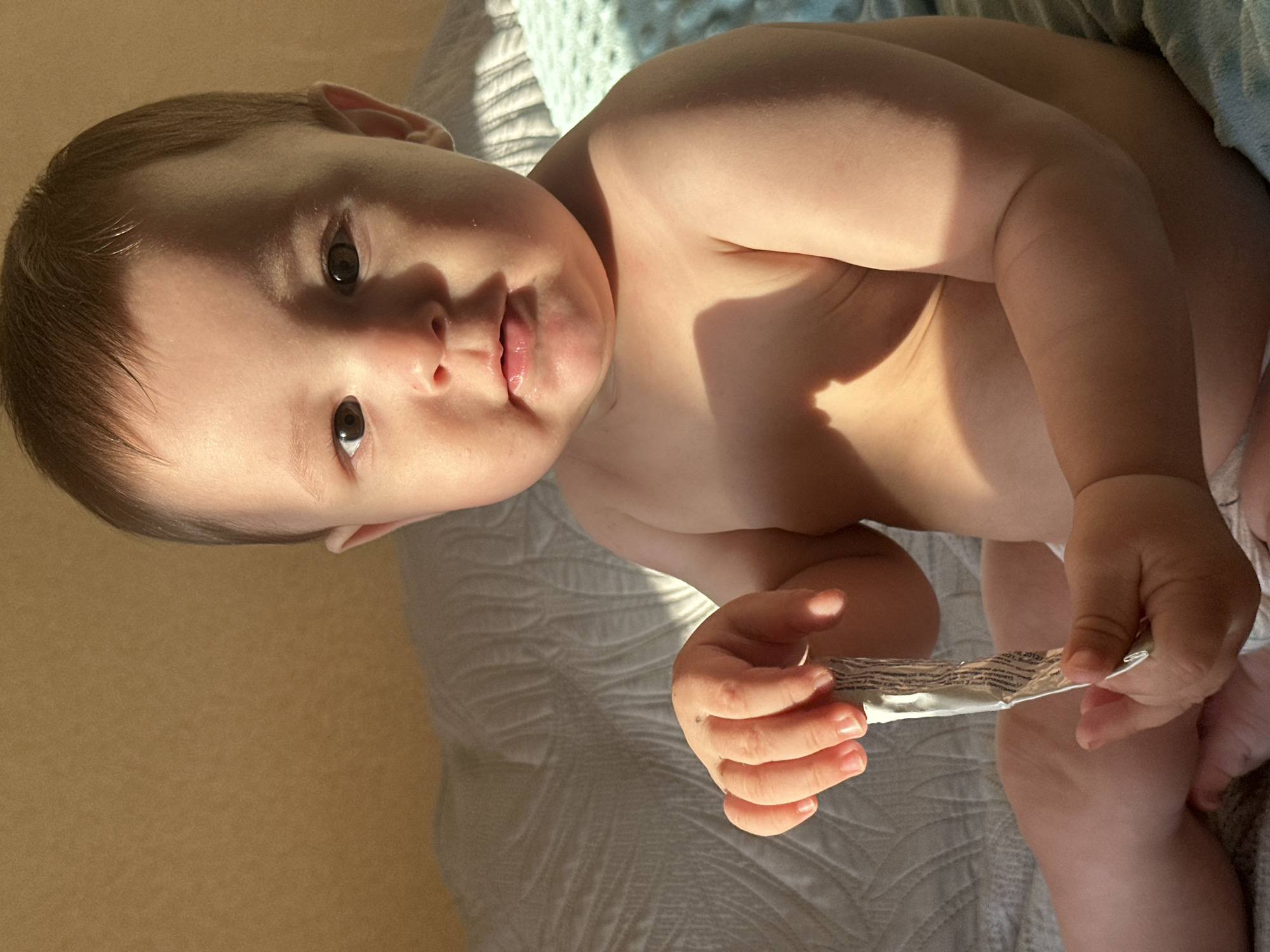 A baby is sitting on a bed with a tube in its hands. The baby is facing the camera, with its head tilted slightly to the left. The baby has dark hair and brown eyes. Its skin is light-skinned. The baby is wearing a diaper. The baby's left hand is holding the tube, and its right hand is also holding the tube. The tube is silver and has some writing on it. The bed is covered with a light-colored blanket with a textured pattern. There is a blue blanket in the background. Sunlight is shining on the baby from the left, creating shadows on its body and the bed. The wall behind the bed is a light brown color.