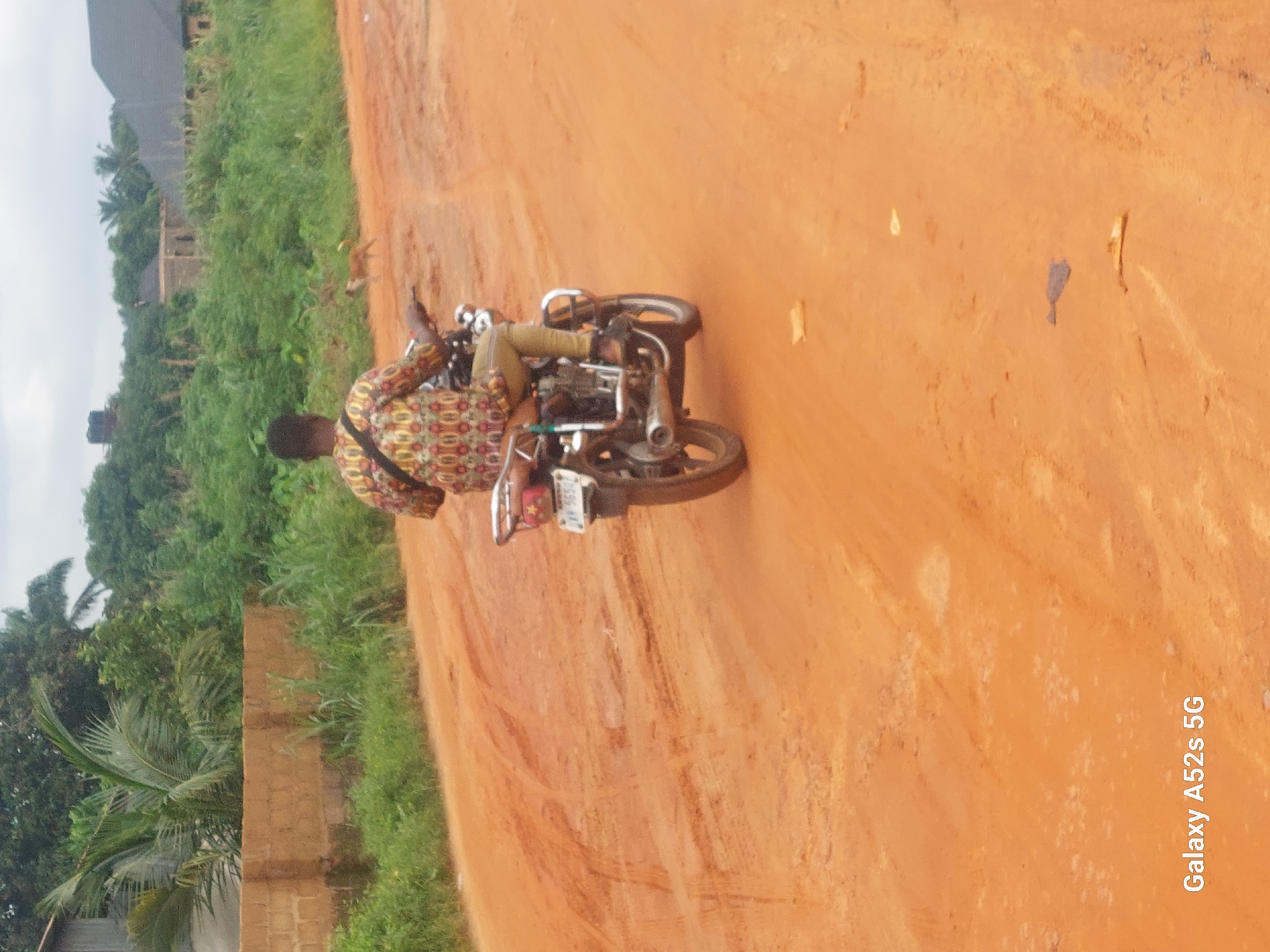 A man rides a motorcycle down a dirt road in Umudioka, Nigeria. The road is reddish-orange, suggesting a laterite soil composition. The man is wearing a colorful, patterned shirt and khaki pants. His motorcycle is dark-colored with a silver exhaust pipe and a license plate that reads "IVM 5650". In the background, there are dense green trees and shrubs, some houses with dark roofs, and a small brown and white dog running. The sky is overcast. The foreground of the image displays tire tracks and debris on the dirt road. The text "Galaxy A52s 5G" is overlaid on the lower left corner of the image, indicating the device used for capture.