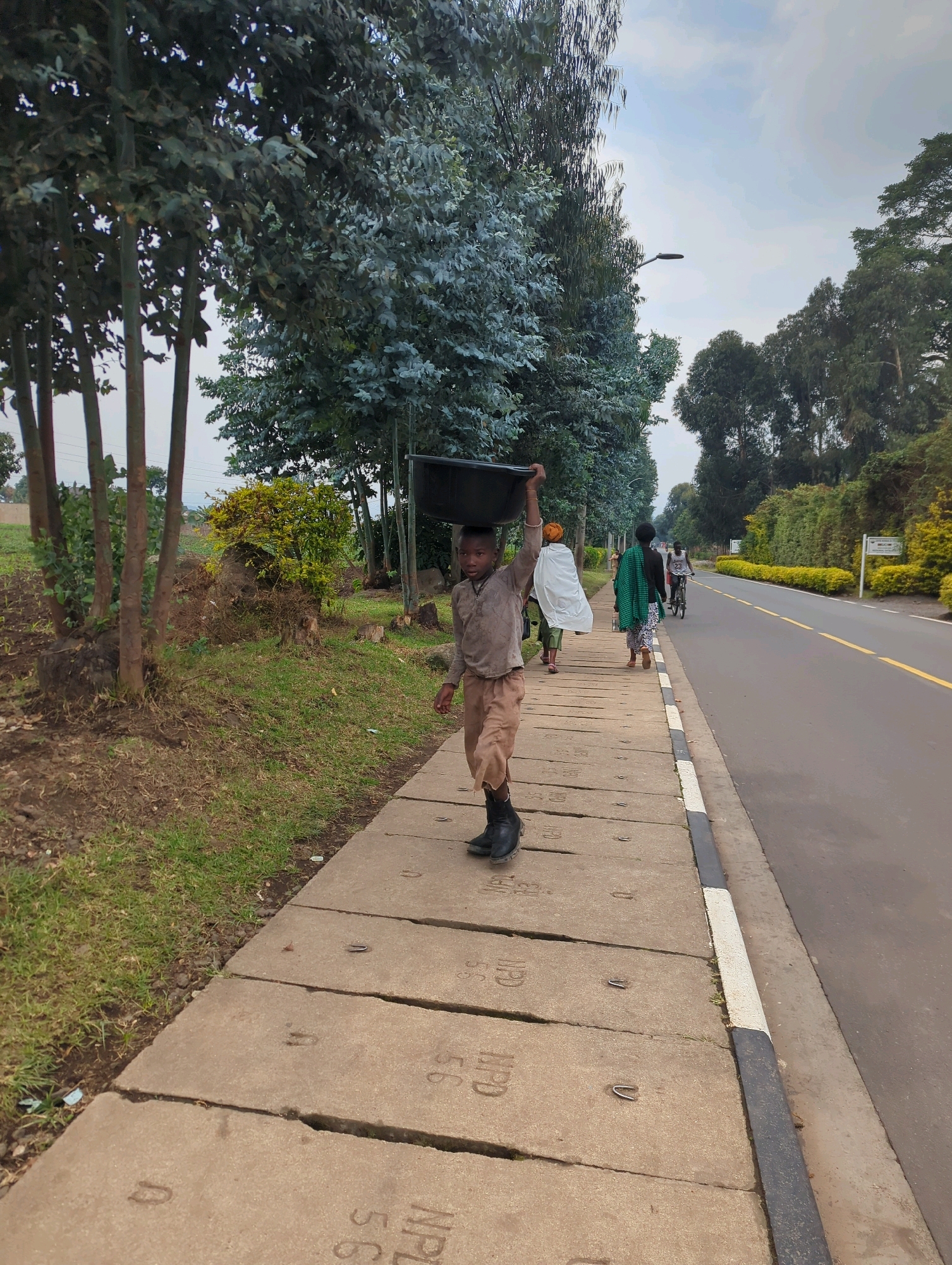 In Musanze District, Rwanda, a young boy walks along a concrete sidewalk, carrying a large, dark basin on his head. He is dressed in a light brown shirt and tan pants, with black boots on his feet. The sidewalk is bordered by a grassy area on one side and a paved road on the other. Several other individuals are visible further down the sidewalk and on the road. One person is riding a bicycle, while others walk. Lush green trees and vegetation line the sides of the road, and the sky is overcast with a light blue hue. The scene suggests a typical day in the area, with people going about their daily activities.