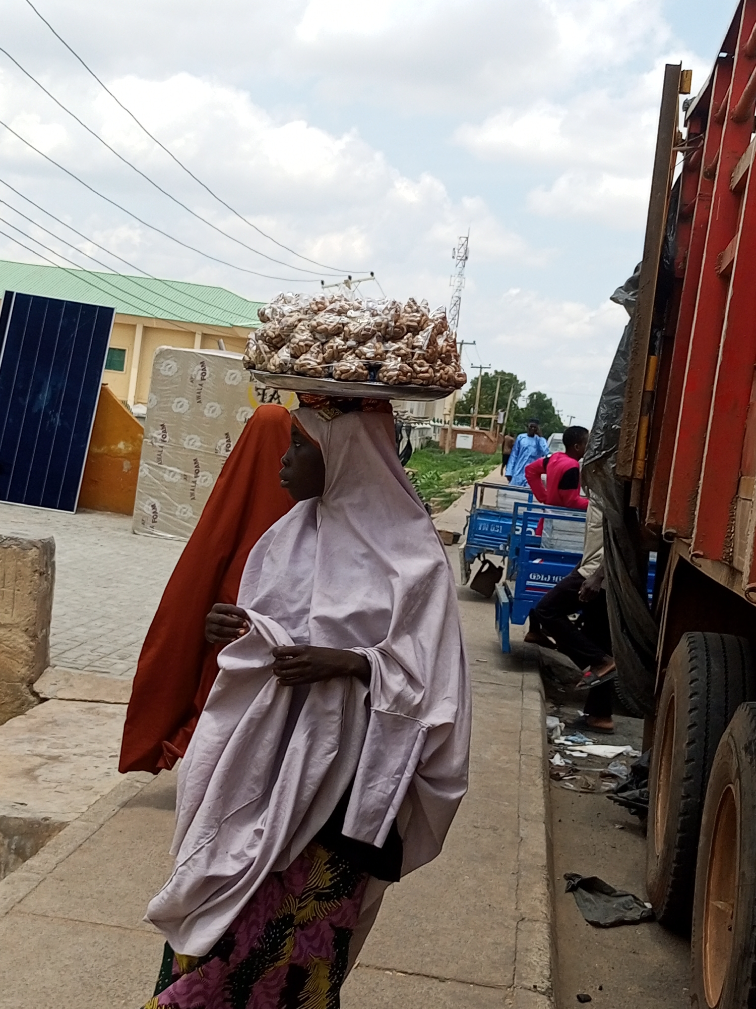 In the city of Arawa, Nigeria, a woman walks along a dusty street, balancing a large tray of goods on her head. She wears a light purple head covering and a dark skirt with a vibrant floral pattern. Beside her, another woman is partially visible, draped in a rust-colored fabric. In the background, a large red truck is parked, its tires and part of its body visible. A small blue cart with two people on it, one in a bright pink top, is positioned near the truck. Further in the distance, a building with a green roof and a solar panel are visible, alongside a few trees. The ground is paved with concrete, and debris litters the curb near the truck. The sky above is cloudy.