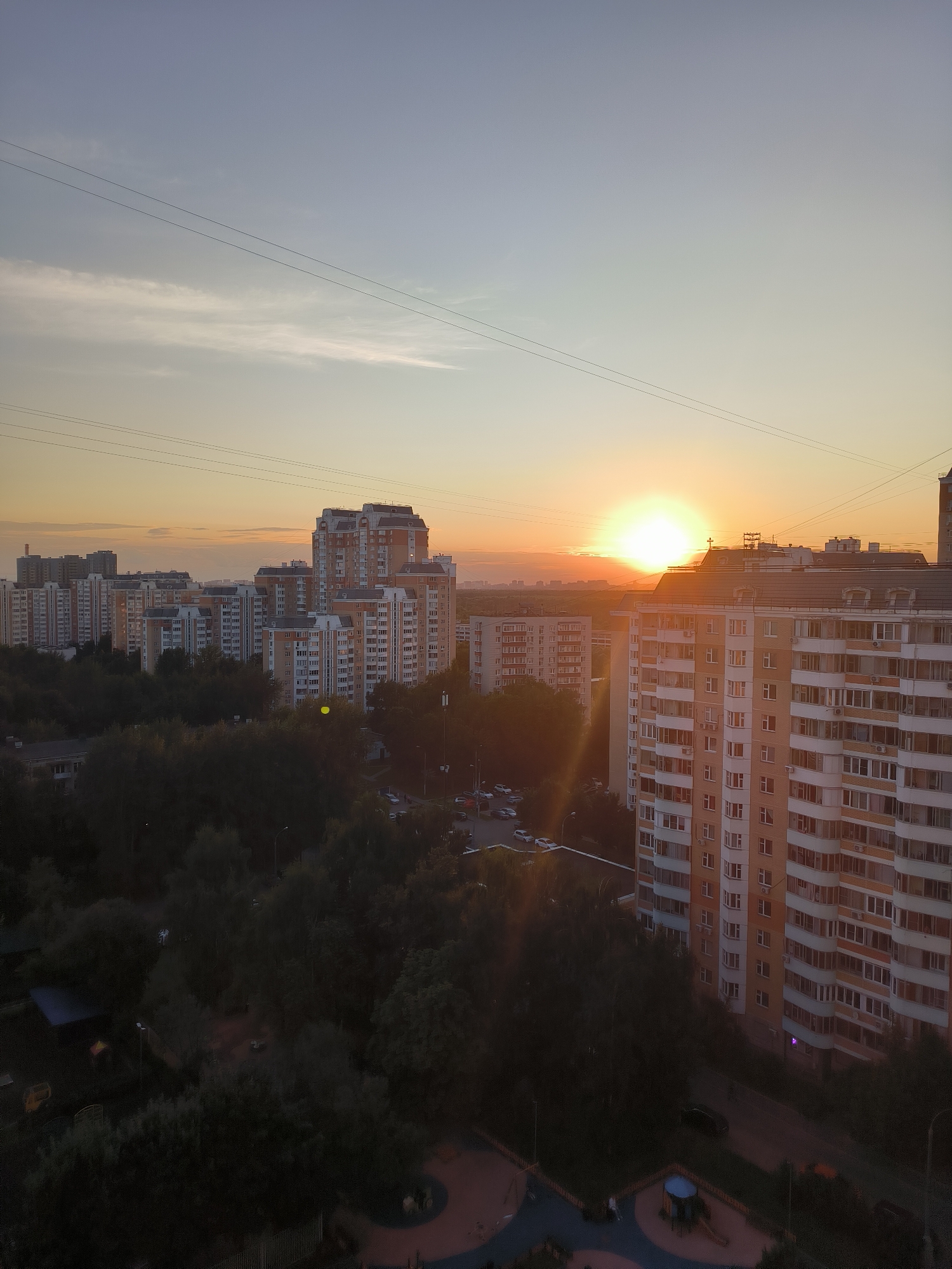 This is a high-angle, wide shot of a residential area in Moscow, Russia, during sunset. The sky is a gradient of light blue and orange, with the sun a bright orb low on the horizon, casting a warm glow over the scene. Several tall, multi-story apartment buildings dominate the midground and foreground. The buildings are a mix of beige, orange, and white, with numerous windows and balconies visible. Trees with dark green foliage form a dense line between the buildings and the foreground. A street with parked cars is visible between the trees and the apartment buildings. In the foreground, there are parts of a children's playground with colorful surfaces and equipment. Overhead, power lines are strung across the sky. The overall atmosphere is one of urban residential life at dusk.
