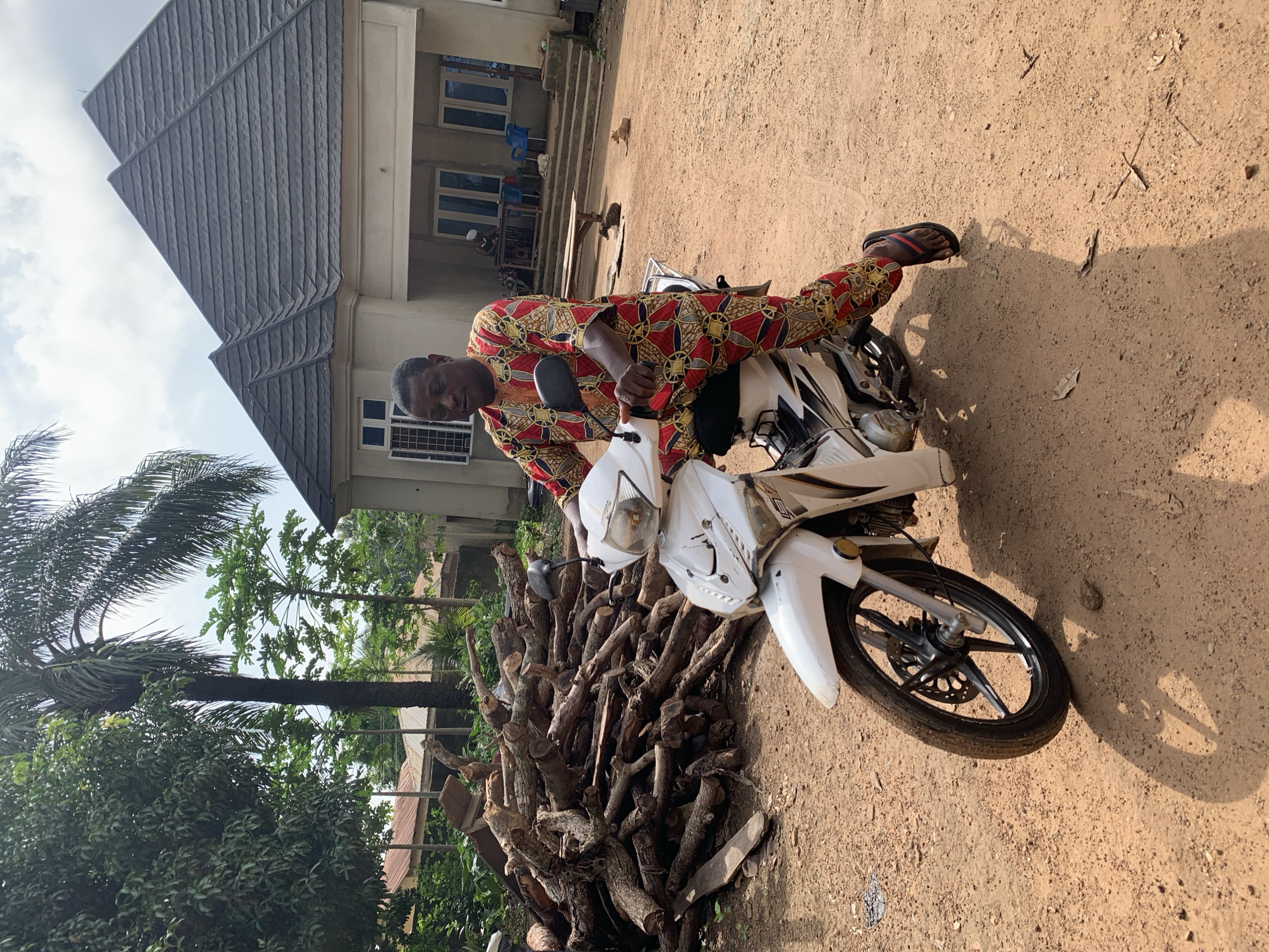 A middle-aged African man wearing a red, yellow, and black patterned traditional outfit is seated on a white motorcycle. The motorcycle is positioned on a dirt path in front of a light-colored building with a dark grey, multi-peaked roof. To the left of the motorcycle and man is a large pile of cut logs. In the background, beyond the building, are more trees and structures. The scene appears to be outdoors in a rural or semi-rural setting. The man is looking towards the right of the frame. Two dogs are visible in the middle distance in front of the building. The lighting suggests it is daytime with some cloud cover. The location is identified as Akwanga, Nigeria.