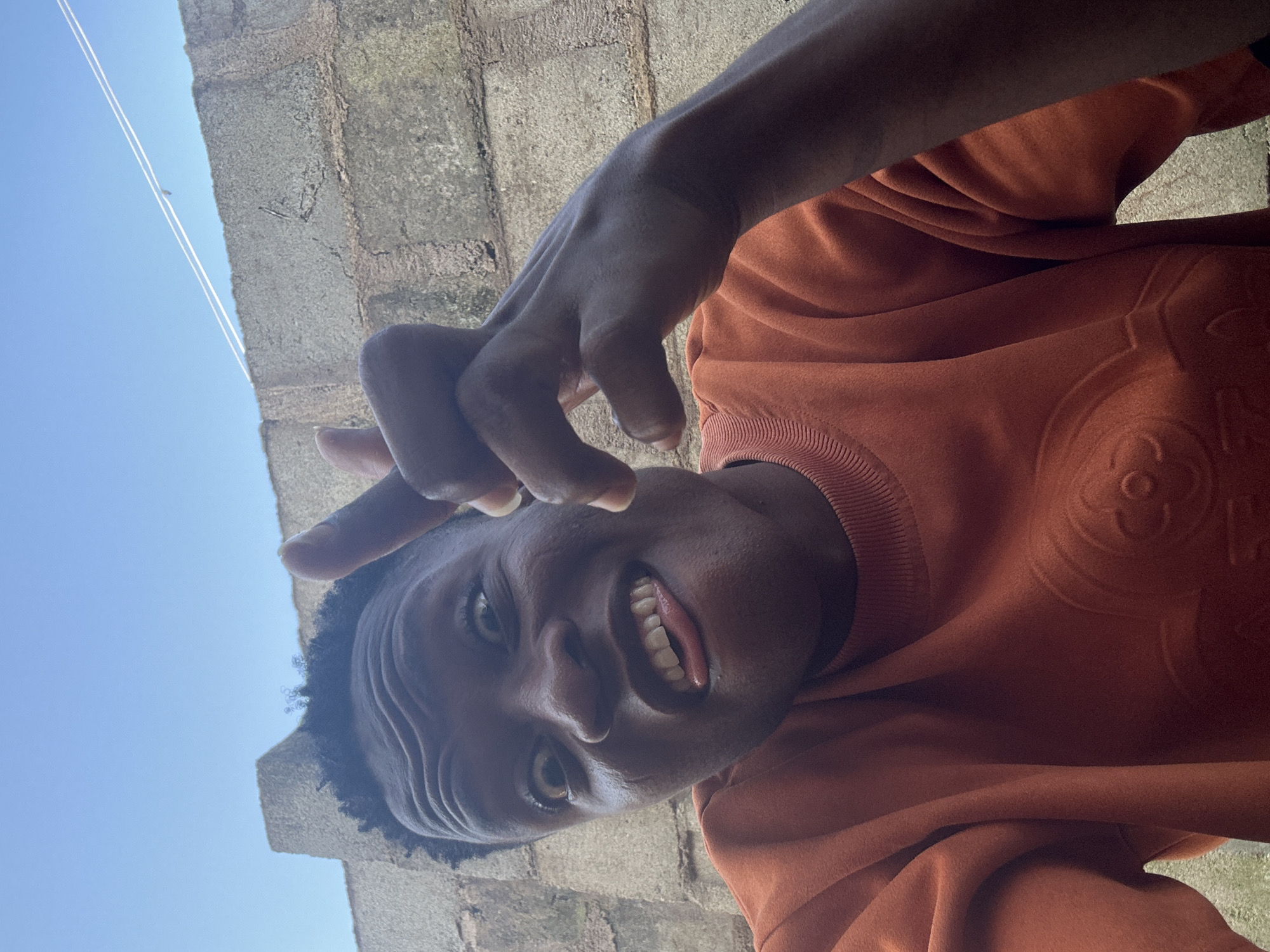 The media file features a close-up, low-angle shot of a young Black person, seemingly a male, outdoors. The subject is looking directly at the camera with a playful expression, sticking out their tongue and smiling widely, revealing white teeth. Their right hand is raised, with fingers curled in a gesture that could be interpreted as playful or informal.
The subject is wearing an orange sweatshirt with a subtle, embossed pattern, possibly a logo, visible on the chest area. The background consists of a brick wall, weathered and textured, and a clear blue sky. Two thin white lines, possibly wires or strings, are visible against the sky in the upper left quadrant of the image. The lighting suggests it is daytime, with bright sunlight casting shadows and illuminating the subject's face.
The overall mood is candid and energetic, captured in a casual outdoor setting. Given the location context of Ikorodu, Nigeria, the scene depicts a moment of everyday life. There is no visible text in the media file.