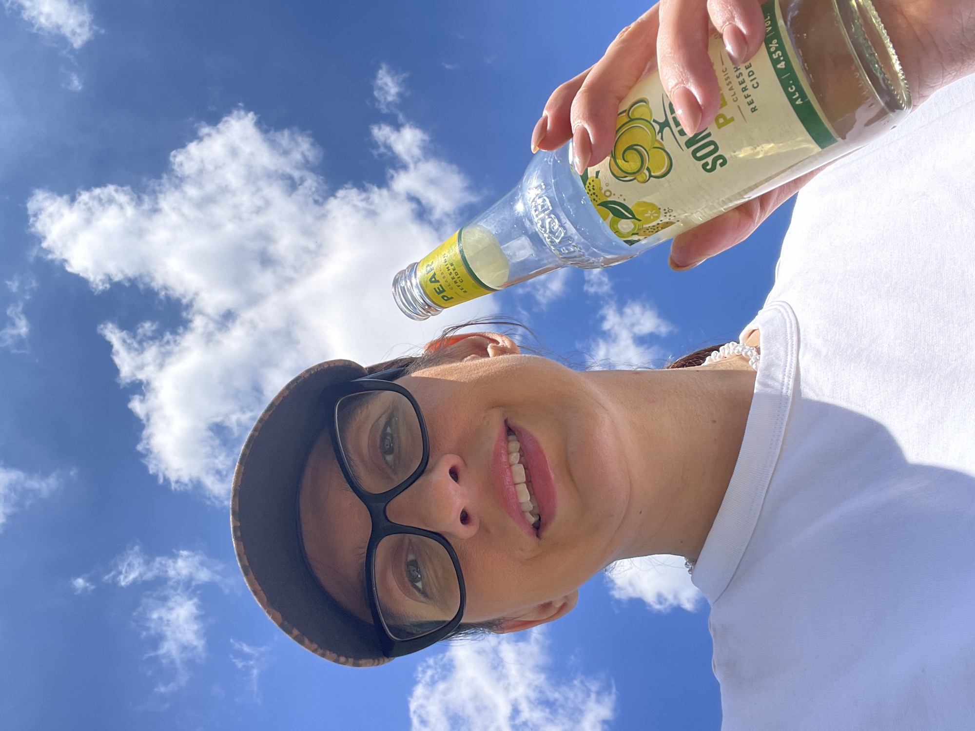 A low-angle shot shows a woman wearing glasses and a hat, holding a bottle of Somersby Pear Cider against a blue sky with fluffy white clouds. She is wearing a white shirt and a pearl necklace. The bottle is angled towards her face as if she is about to drink from it. The bottle label is partially visible, displaying the brand name and "PEAR Classic Refreshing Cider ALC. 4.5% VOL". The background is a bright blue sky with scattered white clouds, suggesting an outdoor location. The exact location in Sofia, Bulgaria is not discernible from the image.
