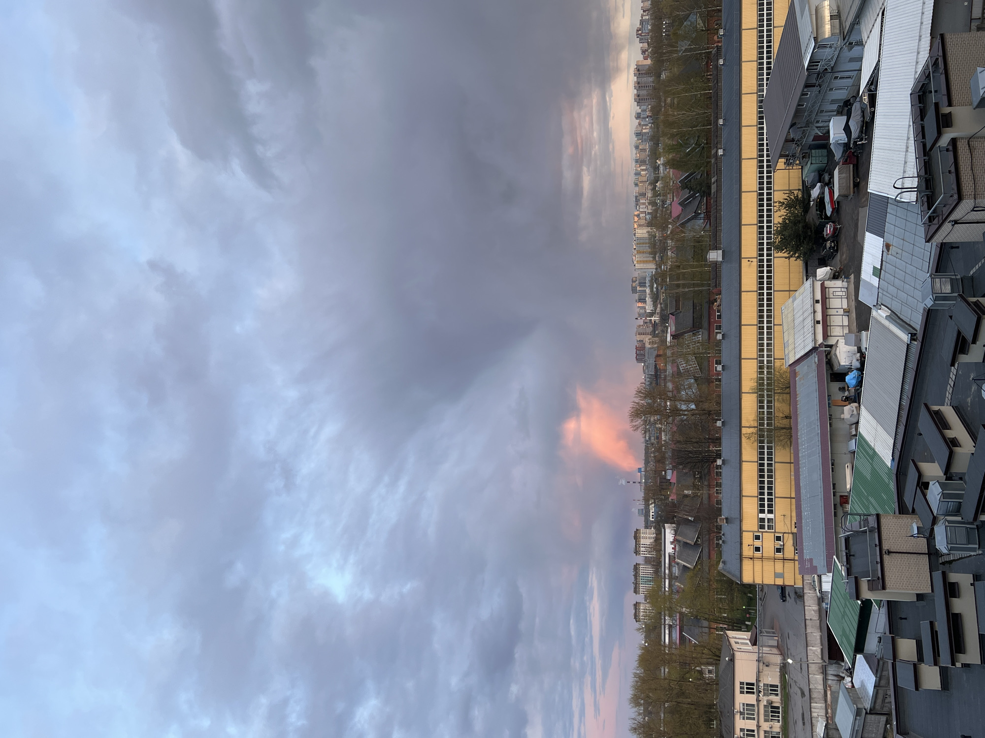 The image depicts an expansive urban and industrial landscape under a heavily clouded sky, viewed from an elevated perspective. The sky is predominantly filled with thick, grey clouds, but patches of lighter blue and soft orange-pink are visible where sunlight penetrates, suggesting either early morning or late afternoon light.
In the foreground and midground, a collection of industrial or commercial buildings is visible. Prominent among these are structures with large, yellow facades punctuated by rows of windows, possibly indicating warehouses or office spaces. Surrounding these are other buildings with varied rooftop designs, featuring grey and green metal sheeting, brickwork, and concrete, often adorned with air conditioning units and vents. A paved yard area below houses several parked vehicles, including what appear to be boats on trailers, alongside various storage containers. A few trees, mostly deciduous with either bare branches or nascent foliage, are scattered within this area.
Further in the background, a sprawling cityscape extends into the distance, characterized by numerous multi-story residential or commercial buildings forming a dense urban horizon. A line of trees creates a natural divider between the closer industrial complex and the distant residential blocks. The overall lighting is diffused by the thick clouds, lending a subdued atmosphere, subtly brightened by the colorful sky streaks. No specific human activity or readable text is discernible. The scene's composition is consistent with a large city like Saint Petersburg.