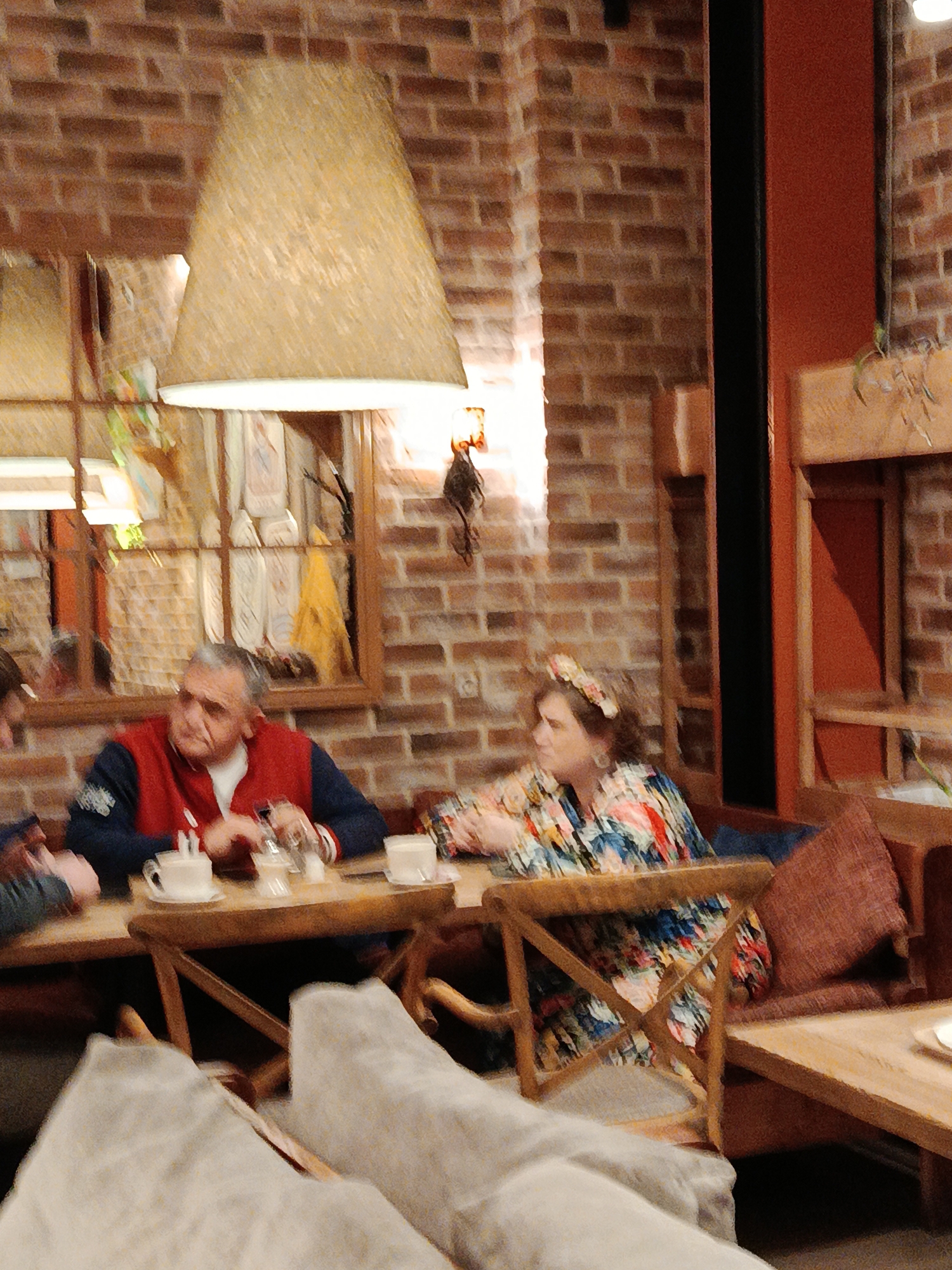 The image shows a brick-walled interior that looks like a cafe or restaurant, likely in Moscow, Russia, given the style of the decor often found in such establishments there. The photo is dimly lit, with a large, conical lampshade hanging from the ceiling. In the background, a mirror reflects the interior.
Two people are seated at a wooden table. On the left is a man wearing a red vest over a dark long-sleeve shirt. He is looking towards the person beside him, who is partially visible. The table in front of him has a coffee cup, a sugar dispenser, and a glass.
To the right is a woman wearing a colorful floral-patterned shirt with a floral headband. She is looking off to the side.
The chairs are wooden with a cross-shaped support in the back. A beige-colored pillow rests on the couch. In the foreground, the back of additional seating is visible.