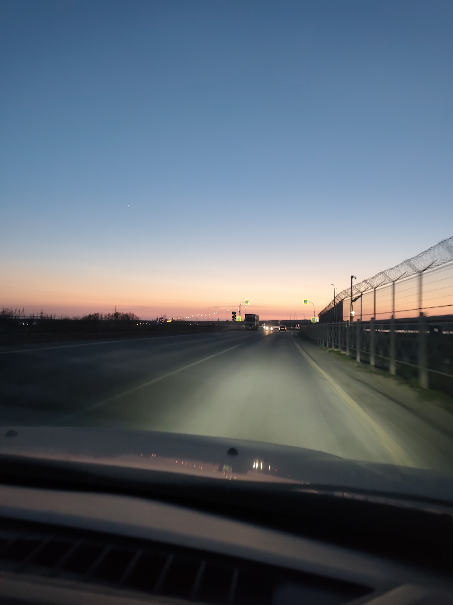 The image depicts a view from inside a vehicle traveling on a multi-lane road towards a sunset. The sky transitions from blue to orange and pink near the horizon. On the right side of the road is a fence topped with barbed wire, possibly indicating a restricted area or perimeter. Several streetlights illuminate the road, and traffic can be seen ahead, including a truck and other vehicles. The location appears to be on the outskirts of a city, with some vegetation and structures visible in the distance. Given the context of Chaikovsky, Russia, it could potentially be a highway leading into or out of the city, possibly near an industrial or security-sensitive site due to the presence of the barbed wire fence.