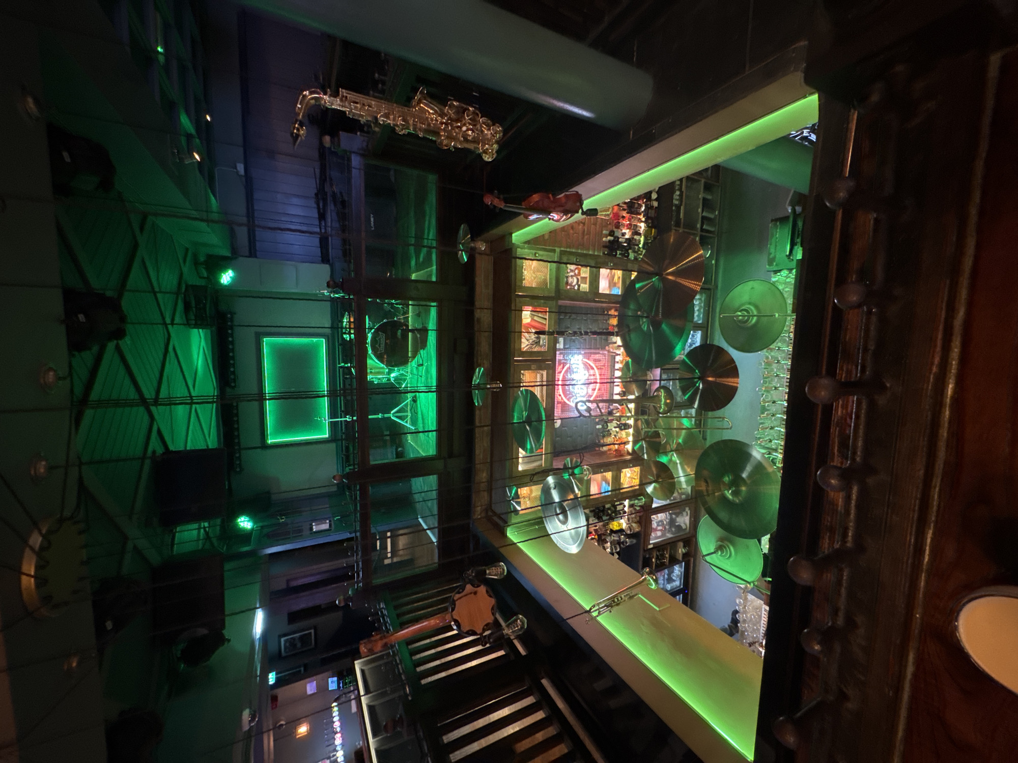 This is an interior shot of a music-themed venue, likely a bar or club, characterized by dramatic green lighting and an array of musical instruments. The image is taken from a high vantage point, looking down into the space.
In the upper central area, a golden saxophone hangs prominently from the ceiling. To its right, an illuminated bar counter features shelves stocked with bottles and decorated with multiple large cymbals. Another stringed instrument, resembling a mandolin or banjo, rests near what appears to be a set of piano keys or a keyboard in the lower left foreground. A drum, possibly part of a drum set, is visible on the left side of the scene, also bathed in green light.
The venue's walls are adorned with various features, including mounted speakers, one of which clearly displays the brand name "LANEY." Geometric patterns and square neon-green light frames contribute to the modern, vibrant atmosphere. A dark wooden railing or banister runs along the right side of the frame, showing decorative metallic knobs. The prevalent green lighting, emanating from LED strips and neon fixtures, casts a strong, almost futuristic glow over the entire space, suggesting an evening or nighttime setting. No people are visible, indicating that no specific event or activity is actively captured, but the setup strongly implies a readiness for live music performances.