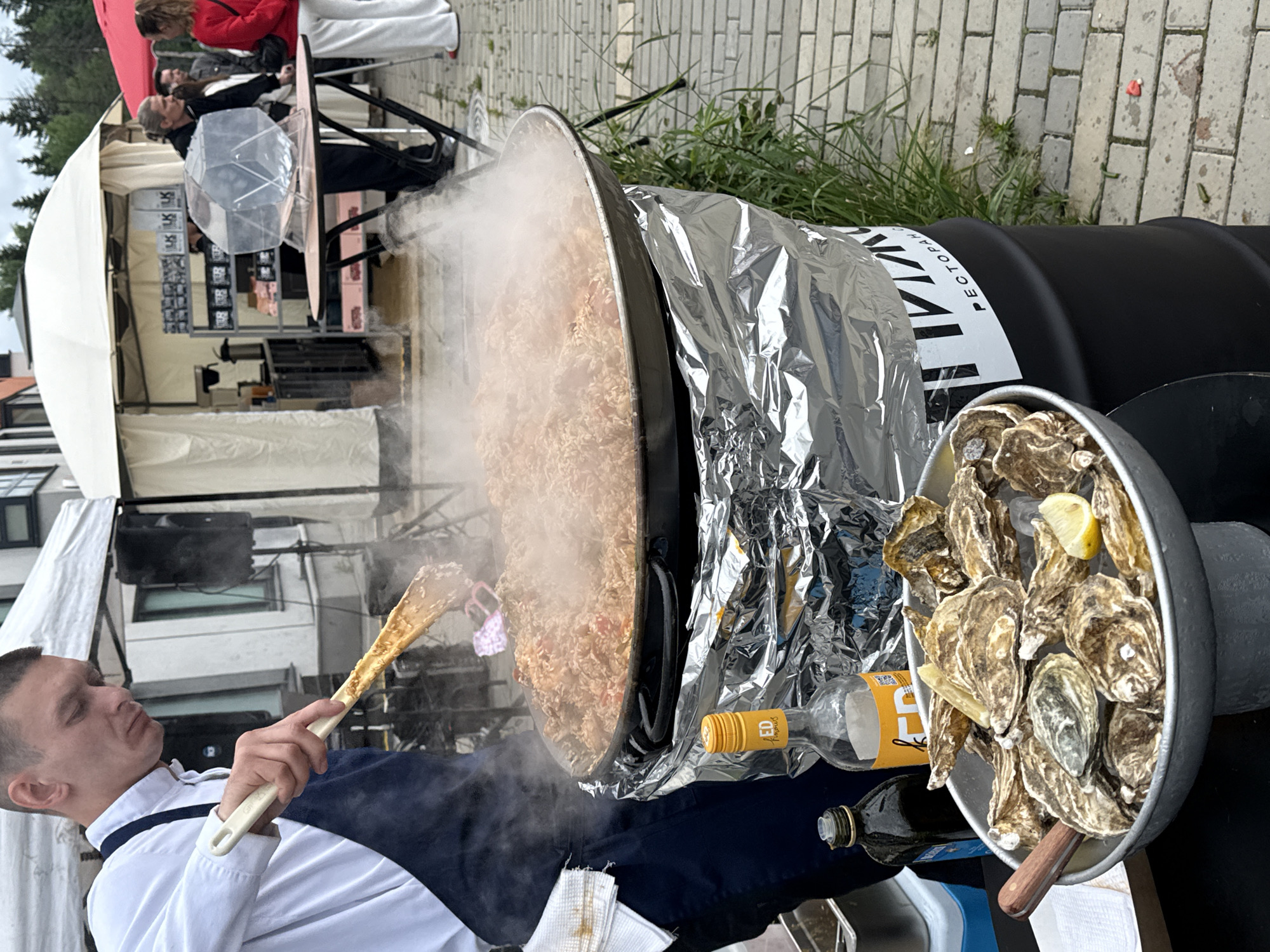 The image depicts an outdoor culinary event in Izhevsk, Russia, under an overcast sky. In the foreground, slightly to the left, a young adult male with short hair, wearing a white collared shirt and a dark blue apron, is actively stirring a large, black, round pan filled with a steaming rice-based dish (resembling paella with visible vegetables and possibly meat). He holds a long-handled light-colored wooden spatula. A white towel is tucked into his apron.
The large cooking pan rests on a black cylindrical barrel, the top of which is covered in wrinkled aluminum foil. Two bottles are visible on the surface near the pan: one dark brown bottle (possibly olive oil) and one clear bottle with a yellow label displaying "ED Famous". The barrel itself has a white label with partial Cyrillic text visible, including "РЕСТОРАН" (RESTORAN).
To the lower left, a large, shallow metal bowl filled with oysters on ice and garnished with lemon wedges is prominently displayed on a dark stand. A small wooden-handled utensil, possibly an oyster knife, is also in the bowl.
In the mid-ground, multiple white canopy tents are set up. One tent, to the right of the cook, features shelves displaying various packaged goods, some marked with "LK". Behind a table in this tent, a man with long hair, dressed in dark clothing, stands near a clear, polygonal container, possibly for a raffle or lottery. He is engaged in conversation with a woman in a red top and white pants, whose back is largely turned to the camera. Another person is partially visible behind them. Further to the right, a red canopy tent is present.
The ground is paved with light-colored bricks or pavers, with some grass and weeds growing in the cracks and along the edges. In the far background, urban buildings are partially visible behind the tents and sparse trees. A black speaker on a stand is visible behind the main cook.