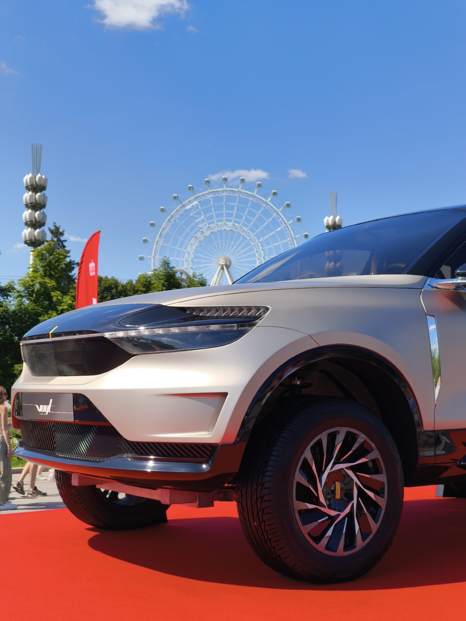 In Moscow, Russia, on a bright sunny day, a modern concept car, likely an SUV with a beige and black exterior, is displayed on a vibrant red carpet. The car features a striking design with sharp lines, sleek headlights, and a prominent grille. Its large, intricately designed wheels are a focal point. In the background, a large Ferris wheel stands tall against a clear blue sky with scattered white clouds. Trees and a red flag are also visible, suggesting an outdoor event or exhibition. A few people are seen walking in the distance, adding a sense of scale and activity to the scene. The image captures a moment of showcasing automotive innovation in an urban, recreational setting.