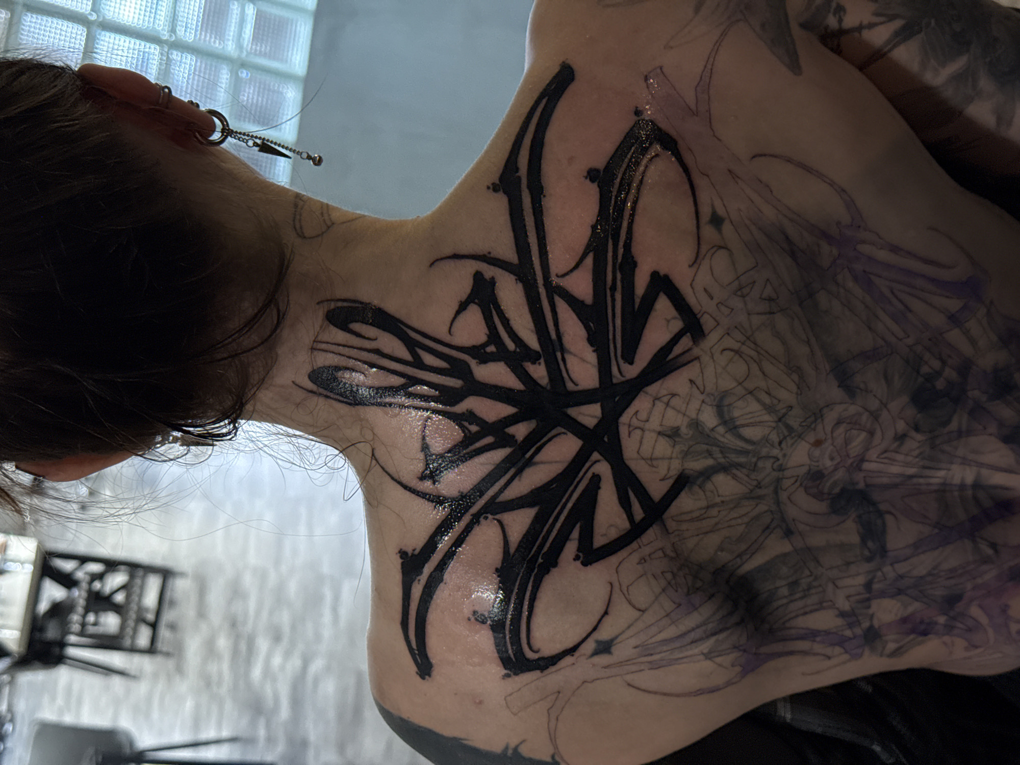 The image shows a person's upper back and neck with fresh tattoos. A large, black, abstract tattoo dominates the upper back, still wet from the tattooing process. Beneath this tattoo, a more faded and intricate tattoo design in lighter ink is visible, covering a larger portion of the back. The person has multiple piercings in their ear with hanging earrings. Hair is dark, and the background is indistinct, showing a wall with some texture and blurred furniture, suggestive of an indoor setting. The image does not contain information about the location in the city of Moscow, Russia.