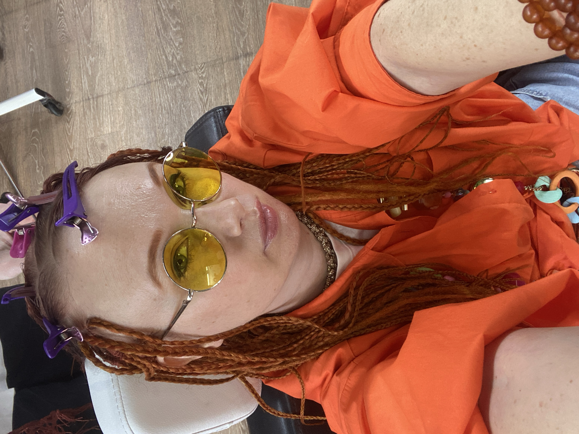An indoor, overhead shot shows a person, likely female, lying down or reclining. The subject has reddish-brown braided hair, possibly dreadlocks, styled with multiple purple and metallic hair clips near the scalp. She is wearing large, round, yellow-tinted sunglasses that reflect a green eye. Her lips are slightly parted, showing a light pink color, and freckles are visible on her skin.
She is dressed in a bright orange oversized shirt or jacket over a white top, with a glimpse of blue jeans on her lower body. Accessories include a gold-toned chain necklace, various colorful beads, and an orange-brown beaded bracelet or necklace. The background features a light wooden floor and parts of a white and black chair or headrest.
The presence of hair clips suggests the person is in the process of, or has recently undergone, a hair styling session. The overall atmosphere appears relaxed, with the subject seemingly posing for the camera. There is no visible text in the image. While the location context is provided as Sochi, Russia, no visual cues within the image confirm this specific geographical setting.