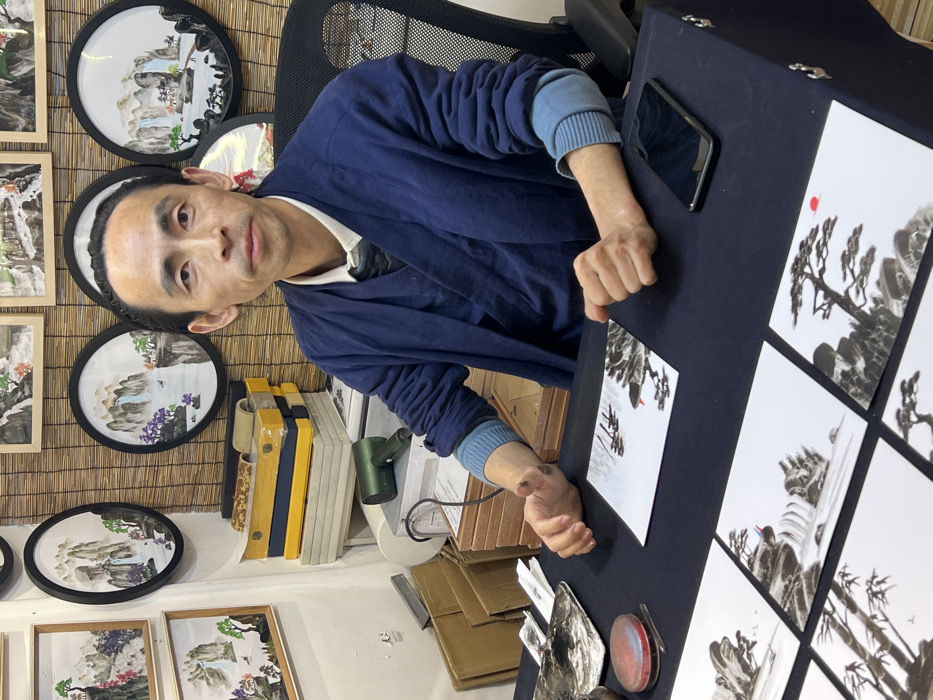 A male individual with dark hair pulled back, wearing a navy blue traditional-style jacket over a lighter blue ribbed long-sleeved shirt and a white collared shirt, sits behind a dark blue tablecloth-covered table. He faces forward, looking directly at the viewer with a neutral expression. His right hand rests flat on the table, and his left hand is positioned above a small, square piece of paper depicting a black ink landscape scene with a red detail. Scattered on the table in front of him are several other similar square pieces of white paper, each featuring distinct black ink wash landscape artworks, some with small red accents. To the left of the man's left hand, there is a metallic ink slab with black residue and a red circular ink pad with a small tool on it. A black smartphone is visible on the table to the right of his right hand.
Behind the man, a bamboo mat-style wall covering is visible. Hung on this wall are multiple framed artworks: several circular frames showcasing landscape paintings with mountains, waterfalls, and trees in black ink and muted colors, and a few rectangular frames with similar themes. To the man's left, a stack of items including yellow boxes, lighter-colored cases, and a green handheld blower are visible on a lower surface. The overall setting appears to be an art studio or shop located in Yuzhong District, China. The man is engaged in, or presenting, traditional Chinese ink painting.