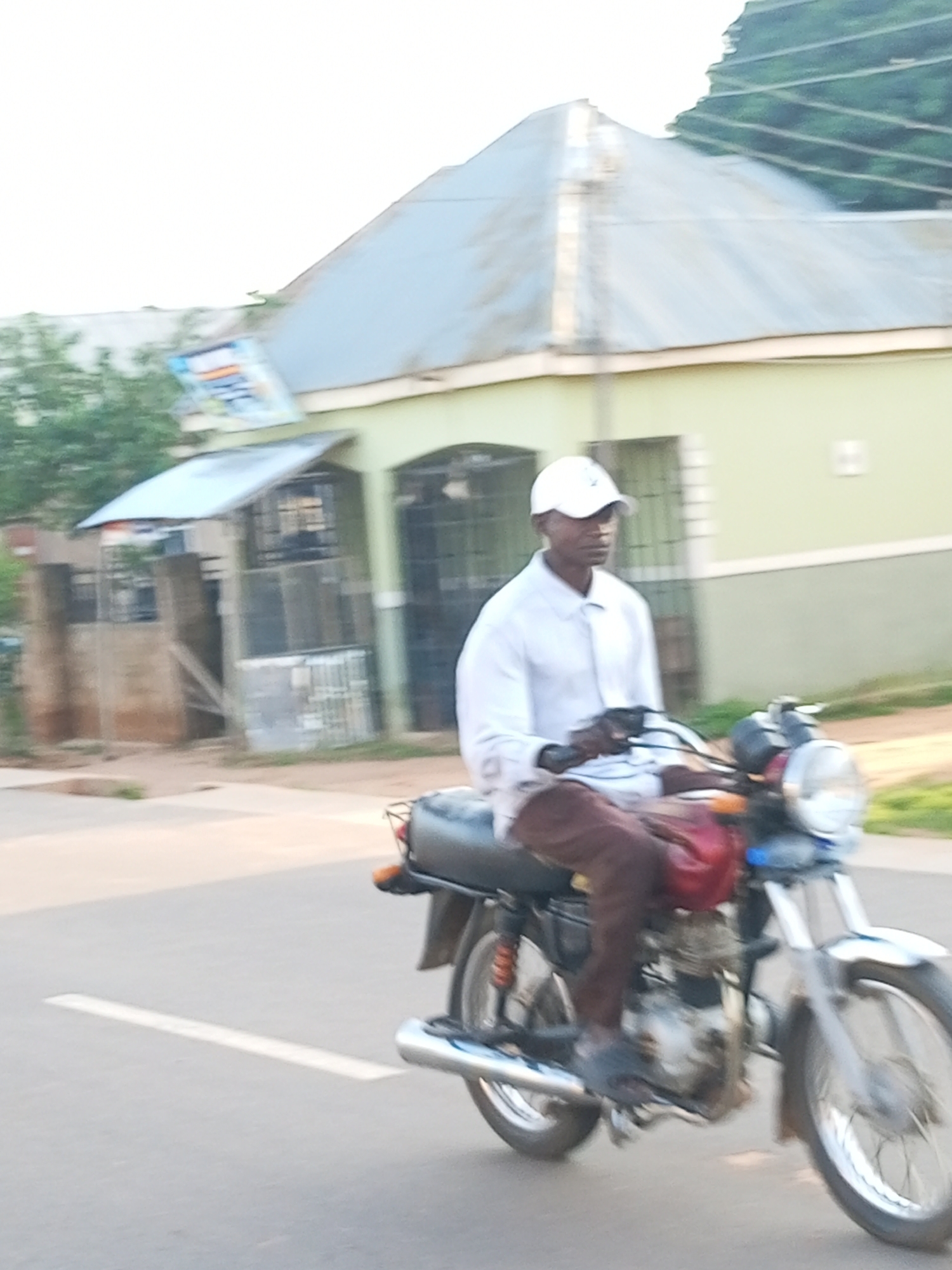 A dark-skinned man wearing a white baseball cap and a white long-sleeved collared shirt is riding a motorcycle on a paved road. He is wearing brown pants and dark shoes. The motorcycle is dark colored with silver accents and a red seat cover. The background shows a light green building with a corrugated metal roof. Trees and a small structure with a metal roof are visible to the left of the building. The image appears to have been taken in Tempure, Nigeria.