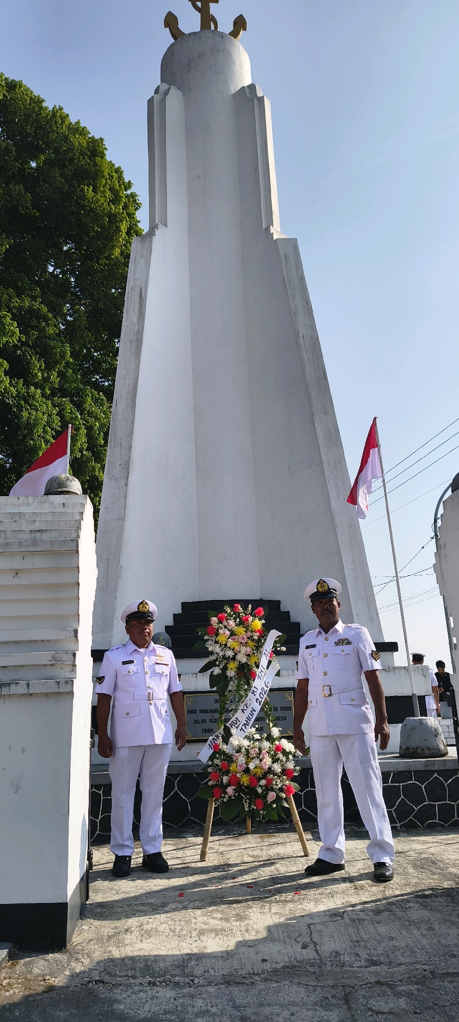 This image captures an outdoor scene in front of a white monument topped with a golden anchor. Two men in white naval uniforms stand symmetrically in the foreground, flanking a floral wreath placed on the ground. The wreath is adorned with a variety of colorful flowers, including red, yellow, and pink, and has a white banner with text. The monument itself is tall and obelisk-shaped, with tiered sections and decorative elements. Red and white Indonesian flags are visible in the background, one on the left and another on the right. The environment appears to be a public space with a clear sky and some trees. The context suggests a memorial service or a ceremony related to Indonesian naval history. The location is identified as Sadamantra, Indonesia.