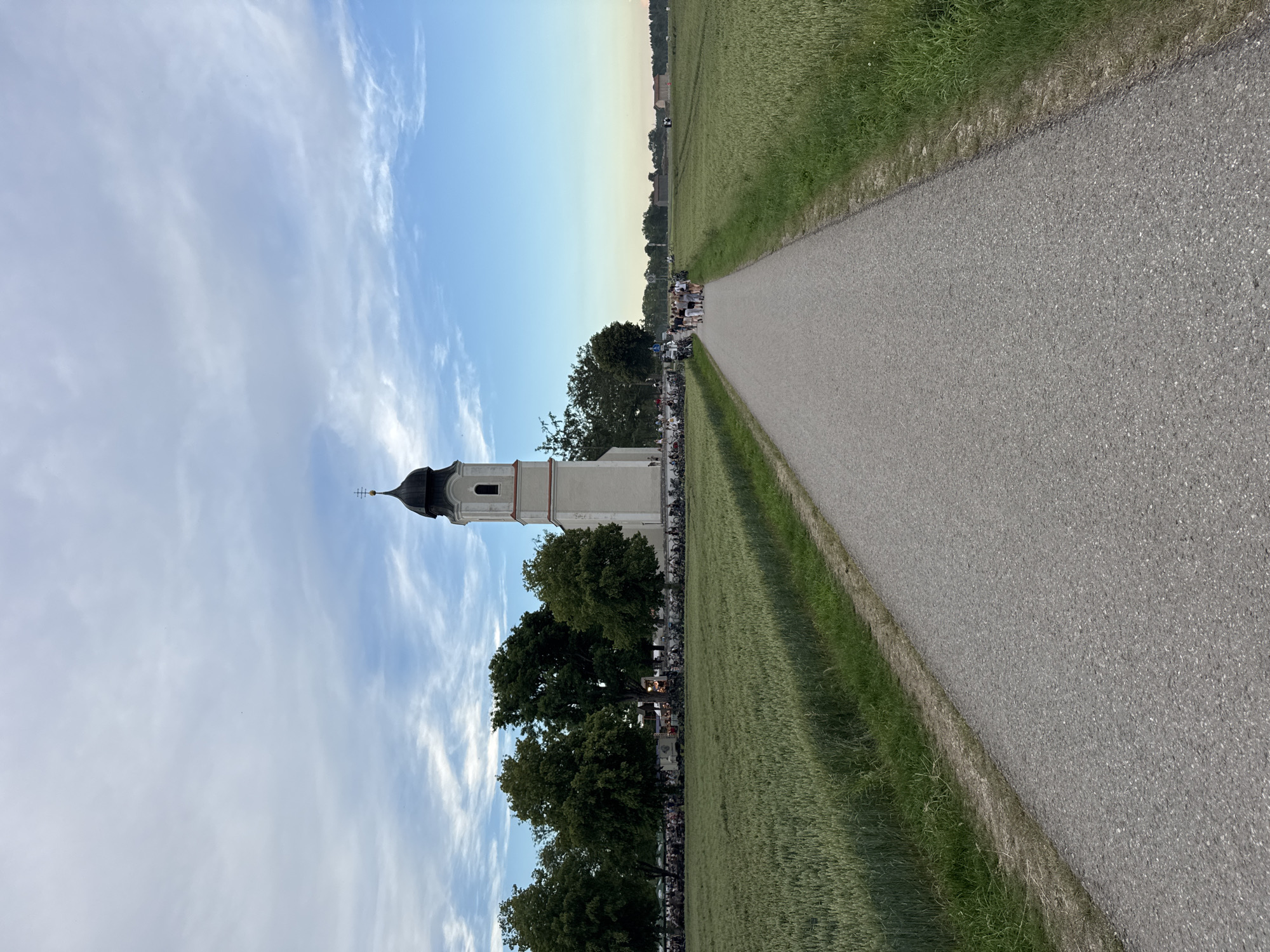 The image depicts a rural scene in Höhenkirchen-Siegertsbrunn, Germany, featuring a white church with a black steeple and a paved pathway leading towards it. The pathway is flanked by fields of green crops on either side. A group of people is visible walking on the pathway, approaching the church, with more people gathered in front of the church and lining the pathway, some appearing to be standing by parked motorcycles. Trees are situated behind the church and to the left of the pathway, and distant buildings and fields are visible in the background under a partly cloudy sky. The overall impression is of an outdoor gathering or event near a church in a rural setting.