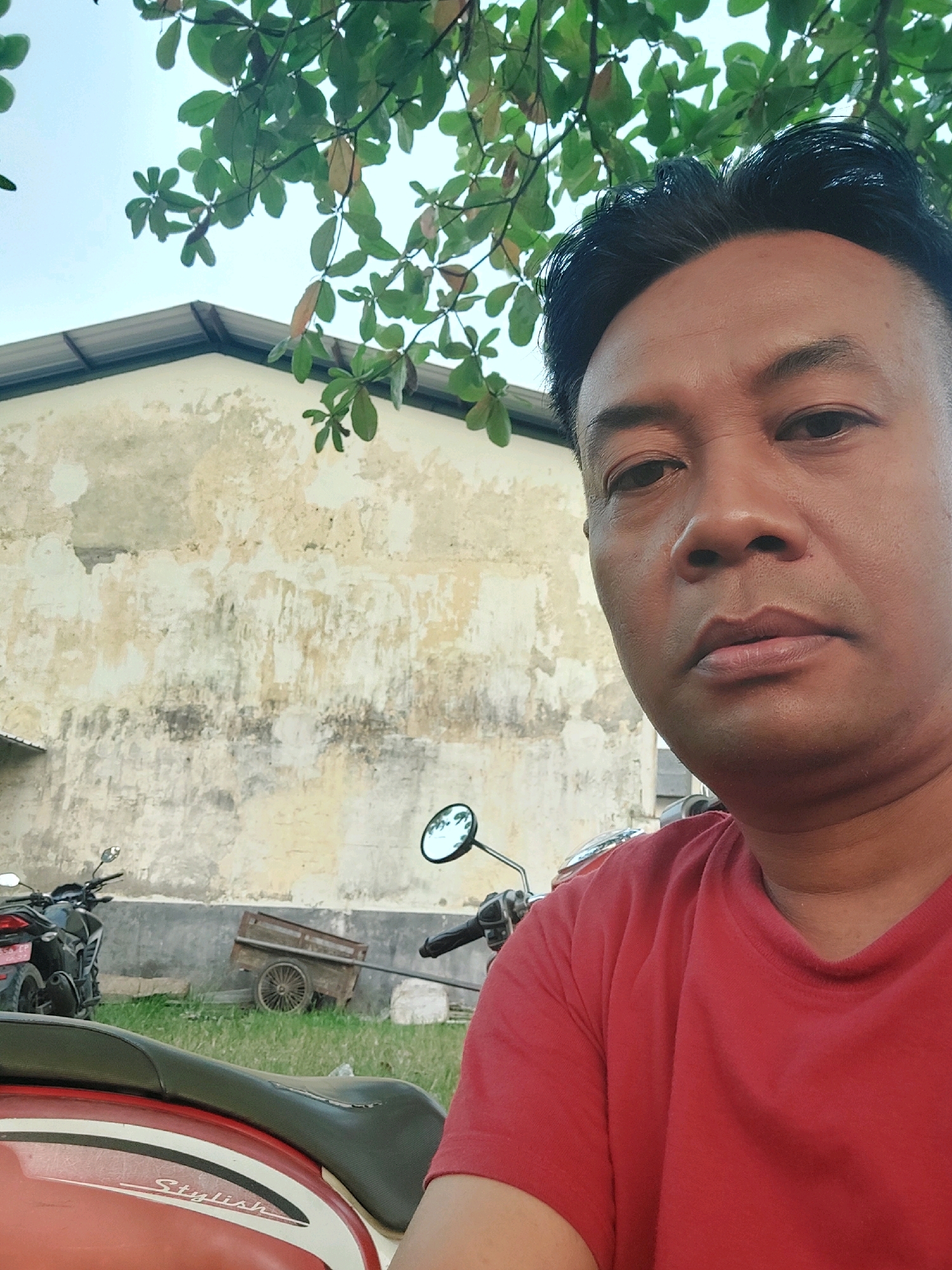In the city of Canga'an, Indonesia, an outdoor selfie is captured. A man with short, dark hair and medium skin tone faces forward, his head and upper torso filling the right side of the frame. He wears a red t-shirt. Behind him, a light-colored, weathered wall with a metal roofline extends across the background. To the left, a black motorcycle is parked on a grassy area. Further left, a small, wooden cart with large spoked wheels is visible. Green leaves from an overhead tree hang into the upper part of the frame. The scene appears to be in a rural or semi-urban setting, likely a yard or alleyway.