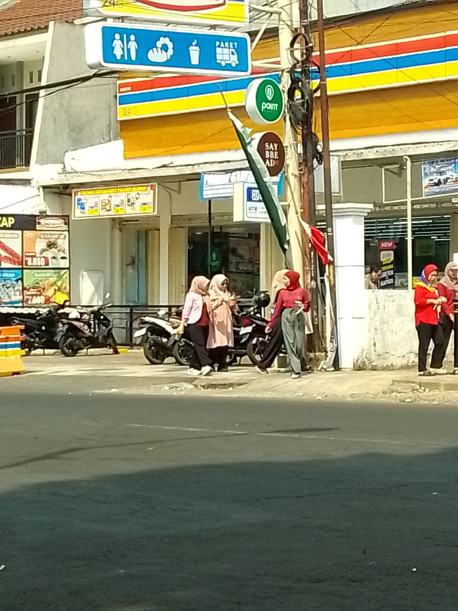 The image shows a street scene in Jember, Indonesia, likely in front of a convenience store or minimarket. The building in the background features multiple signs with Indonesian text and logos, including a blue sign with icons of people, bread, and a cup, and a red, blue, and yellow striped awning. A green circular sign with a white "point" logo is visible on a pole. Several motorbikes are parked along the sidewalk. Three women wearing headscarves are standing near the motorbikes, interacting with each other. One woman in a pink headscarf and light-colored dress stands next to another woman in a similar outfit. A third woman in a red top and grey pants is walking away from them. Further down the sidewalk, two other women in headscarves are also visible, one wearing a red top and the other a light-colored headscarf and pink top. The street in the foreground is paved and appears to be a road with a slight incline. The overall atmosphere is that of a typical daytime scene in an Indonesian urban or semi-urban environment.