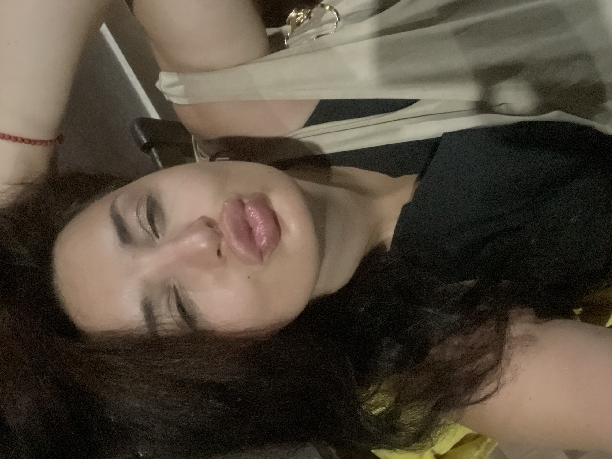 A close-up selfie of a woman with dark hair, pouting her lips, is captured indoors. She is wearing a black top and a cream-colored bag is draped over her shoulder. A red beaded bracelet is visible on her wrist. The background suggests an indoor setting with dark furniture. The lighting is dim. There is no information in the image to suggest the location is Sokhumi, Abkhazia.