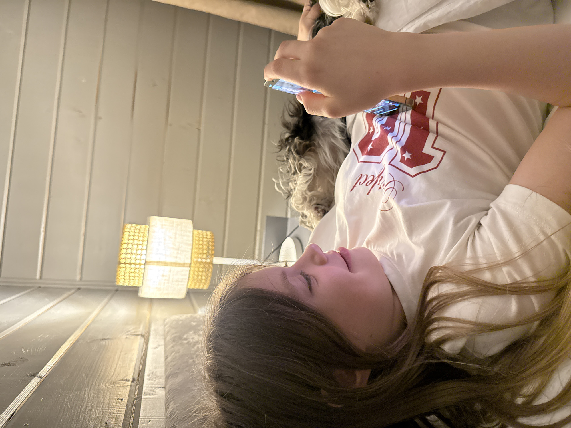 A young person, likely a child with long brown hair, is lying down and looking at a smartphone. The person is wearing a white t-shirt with a red and white graphic. A small dog with black and white fur is visible near the child's head. The background shows a light-colored wall and a lamp. The floor appears to be made of wooden planks. Location: Buzhaninovo, Russia, cannot be determined from the image.