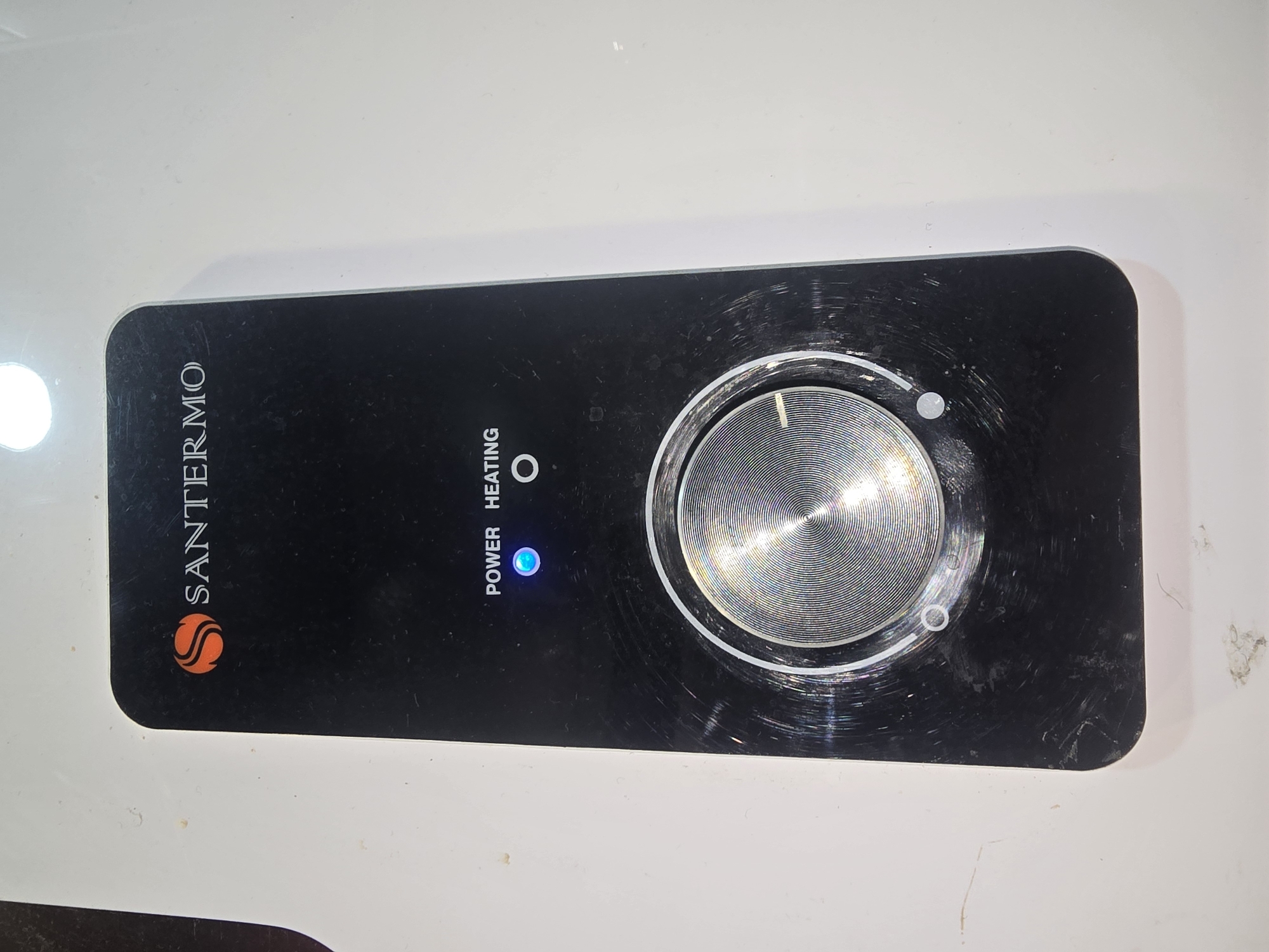 The image shows a close-up of a black control panel with a silver rotary dial. The brand "SANTERMO" is visible at the top of the panel, with an orange and white logo. Below the brand name, the words "POWER" and "HEATING" are printed, with a blue LED light illuminated next to "POWER" and a circular icon next to "HEATING". The rotary dial has concentric circles and a white indicator line. The panel is mounted on a white surface, possibly a wall or appliance. There are scratches and smudges on the black panel. The environment is not discernible beyond the immediate object and its mounting surface. There are no people or animals present. The image does not provide any information about the location in the city of Ulan-Ude, Russia.