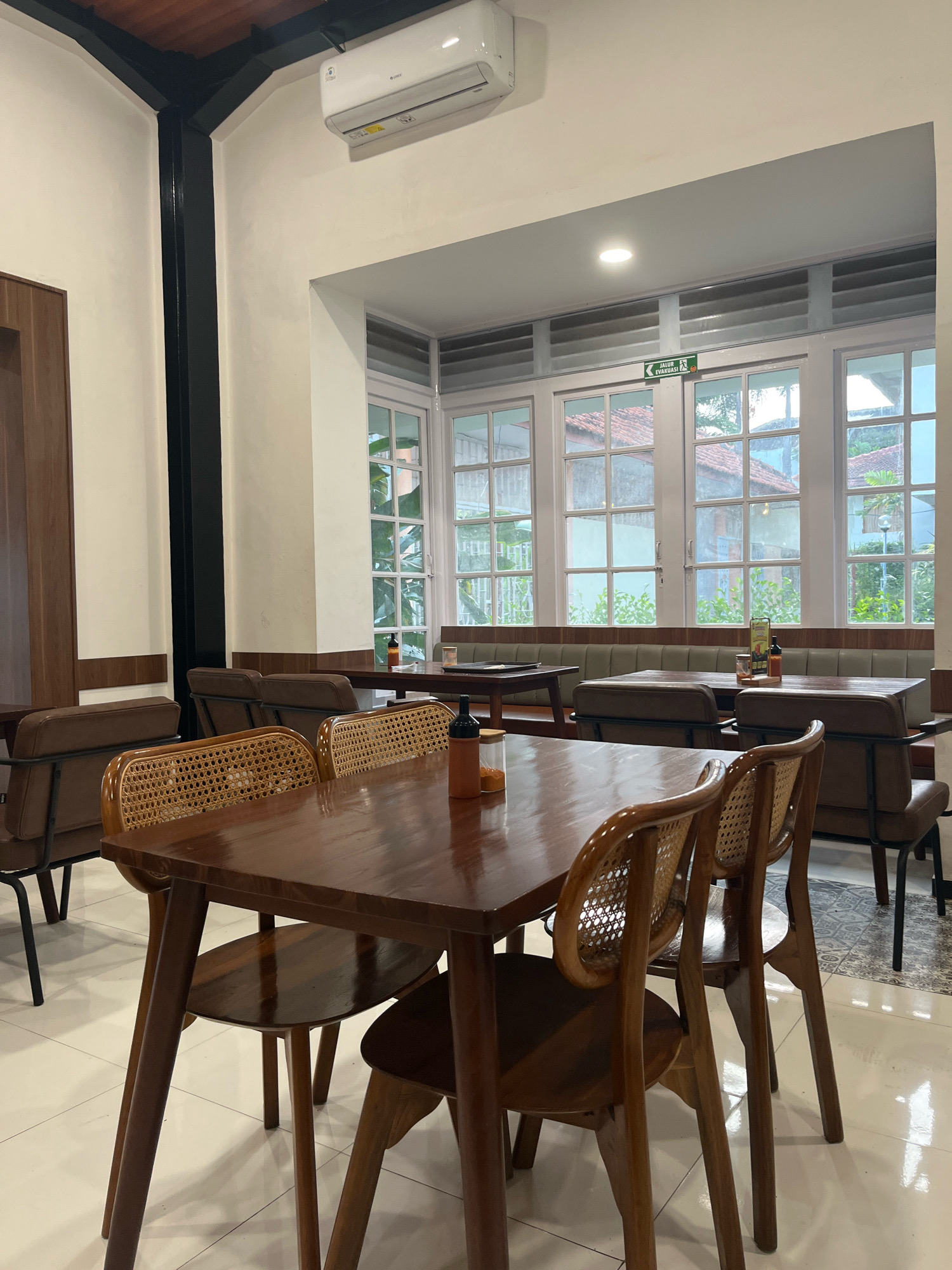 This is a photograph of the interior of a cafe located in Semarang, Indonesia. The cafe features a warm and inviting ambiance with wooden furniture and ample natural light. In the foreground, a dark wooden table is set with two chairs, one with a woven rattan back and the other a fully upholstered brown chair. Two bottles of sauce are placed on the table. In the midground, a long banquette seat runs along a wall with windows. Two wooden tables are visible in front of the banquette, each with chairs and sauce bottles. The windows in the background are large and divided into panes, offering a view of greenery and buildings outside. An "EVACUATION ROUTE" sign is visible above the windows. The floor is tiled with a white and grey geometric pattern. An air conditioning unit is mounted on the wall near the ceiling. The overall scene depicts a well-maintained and modern cafe space, likely designed for casual dining.