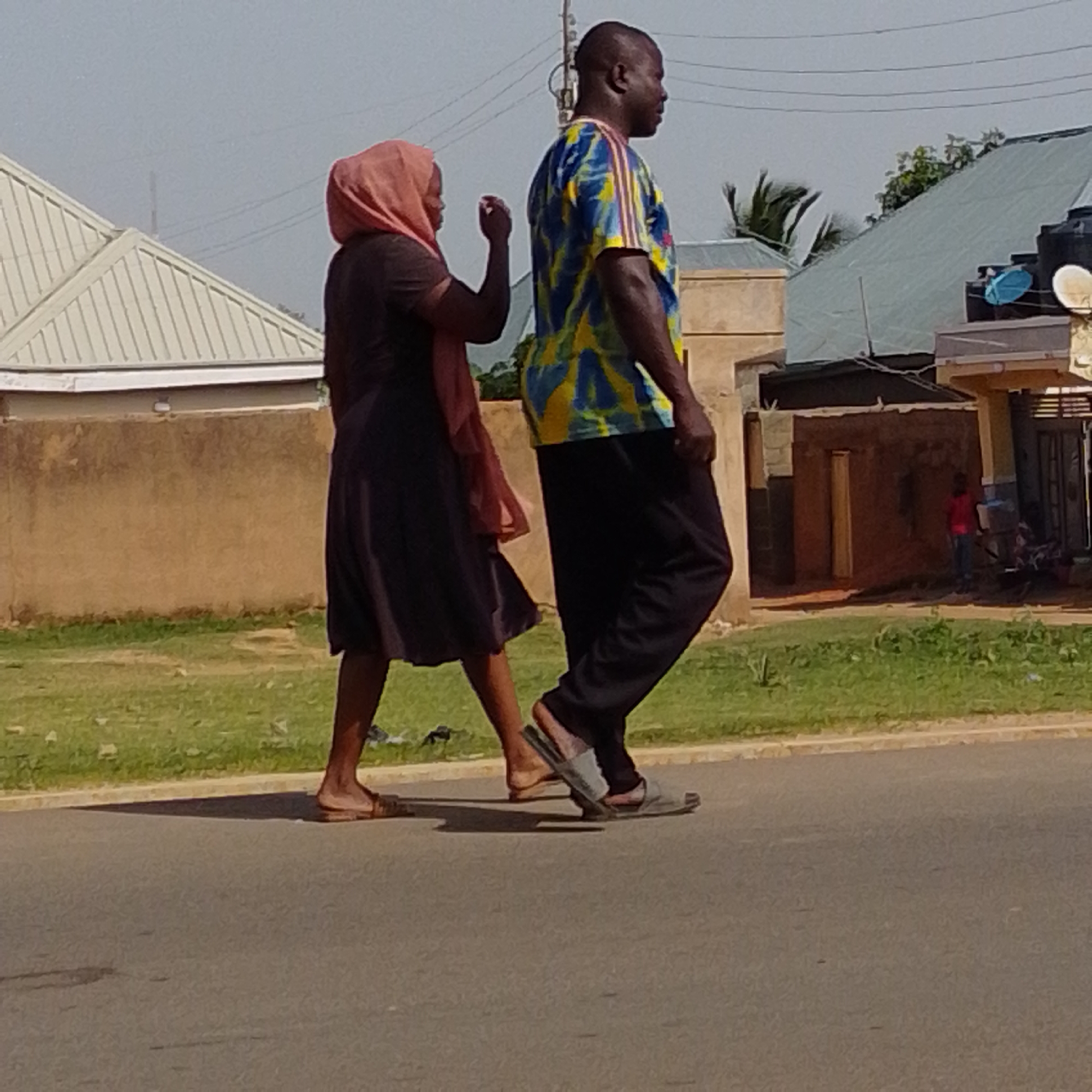 A man and a woman are walking on a paved road. The woman is on the left, wearing a dark dress and a pink headscarf. The man is on the right, wearing a colorful tie-dye t-shirt and dark pants. They appear to be walking in the same direction, with the man slightly ahead. The road is a light gray asphalt. To the left of the road is a strip of green grass. Behind the grass, there is a brown textured wall, and behind that, a light-colored structure with a sloped roof. To the right of the road, there is more green grass and further in the background, buildings with various roof types, including a green metal roof and a dark water tank. The sky is a pale blue with some haze. The scene appears to be in an outdoor, possibly urban or semi-urban environment, consistent with a location in Gombe, Nigeria.