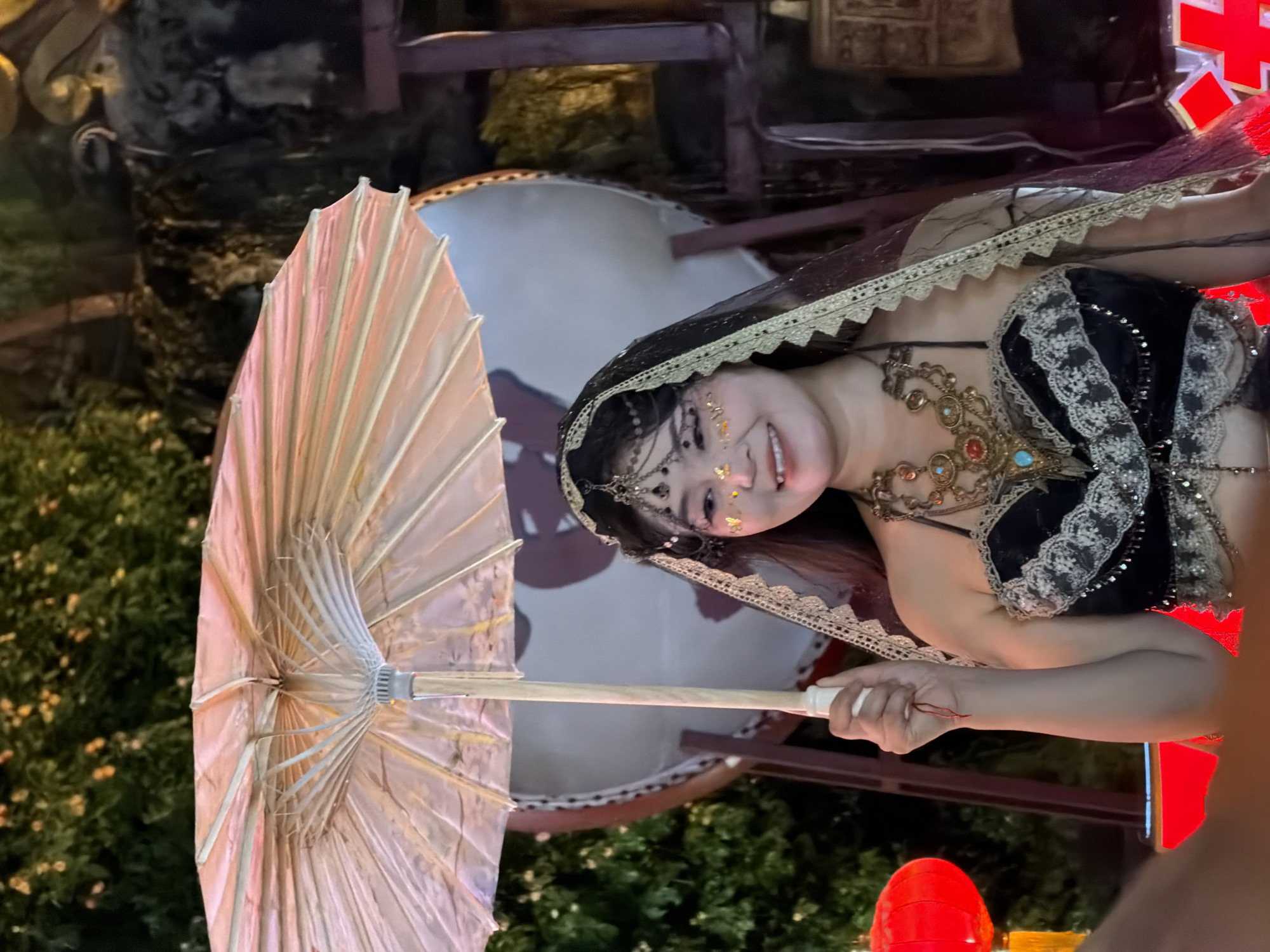 A woman in a black and gold costume stands outdoors, holding a light-colored parasol over her head. She wears a veil with decorative trim, a large ornate necklace, and what appears to be body glitter on her face. Behind her, a large drum is visible, as well as greenery and what looks like a rock formation. A red lantern is also present on the left side of the frame. The overall scene suggests a cultural performance or festival.