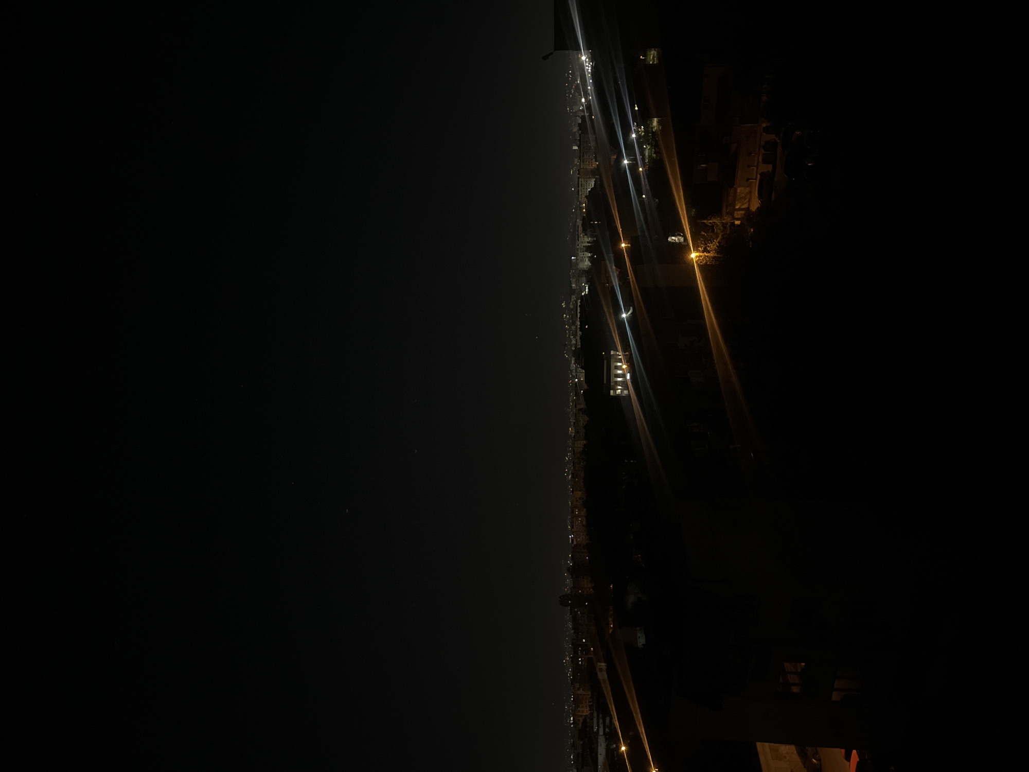 An expansive, wide-angle shot captures a night view of Sofia, Bulgaria. The image is horizontally composed, with a distinct horizon line dividing it. The right half of the frame reveals a sprawling cityscape, illuminated by numerous artificial lights. These lights, ranging from bright white to warm yellow, emanate from buildings, streets, and possibly vehicles, creating a vibrant, glowing line across the distant urban landscape. Some light sources exhibit prominent lens flares or streaks, indicating either a long exposure or specific lens characteristics. Silhouettes of buildings are faintly discernible against the dark backdrop. The left half of the image is predominantly dark, representing the night sky, with a few minuscule, faint points of light that could be stars or very distant, sparse illumination. There is no visible activity, people, or discernible text. The scene conveys a sense of peaceful urban vastness during nighttime hours, under clear weather conditions.