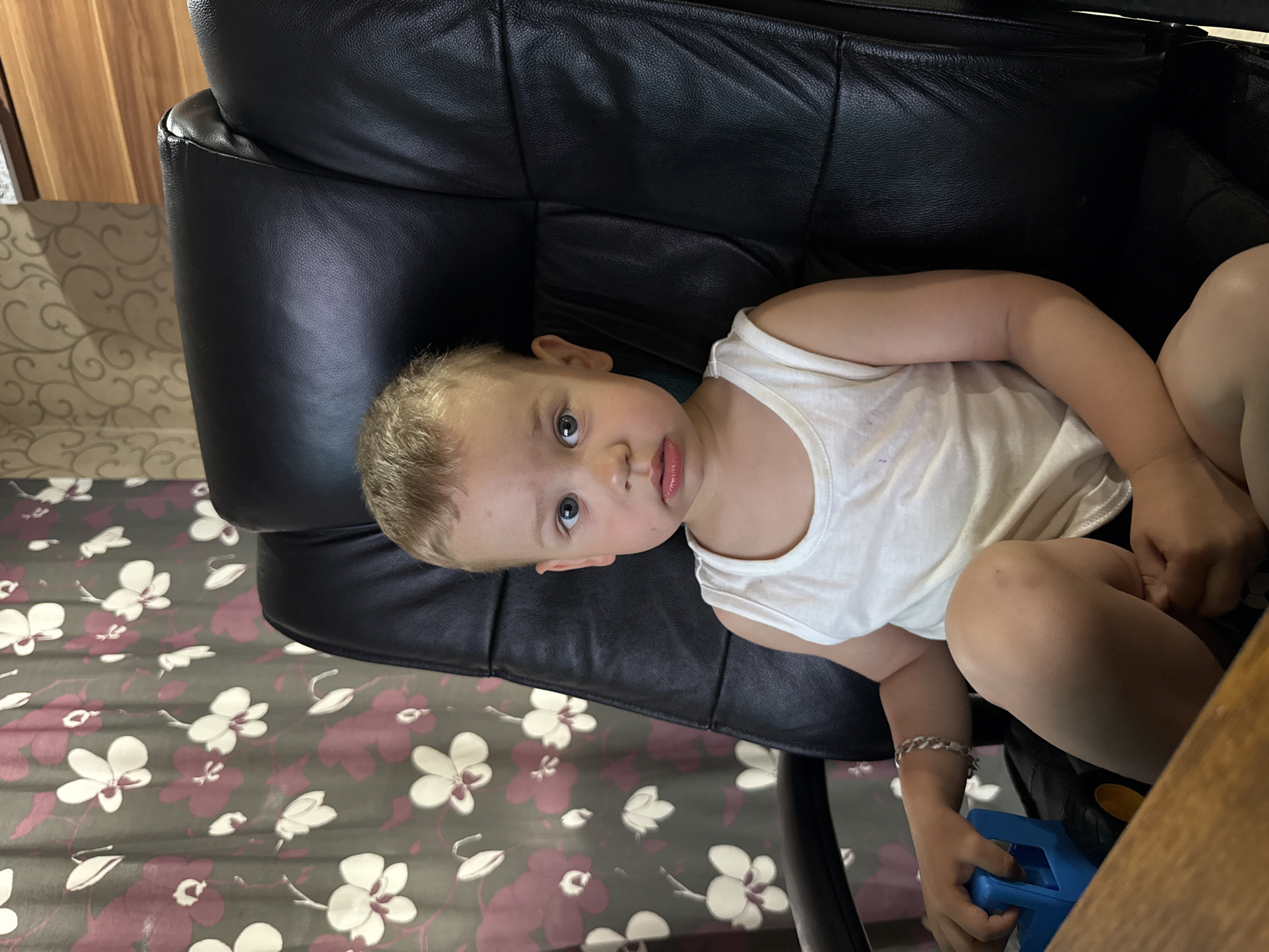 A young child is lying in a black leather chair, facing the camera with a slightly sullen expression. The child is wearing a white tank top. The chair sits on a patterned floor with a floral design in shades of gray, white, and maroon. The edge of a wooden table is visible in the lower right corner of the image. The location is potentially Rusanovo, Russia, but there is no visual confirmation.